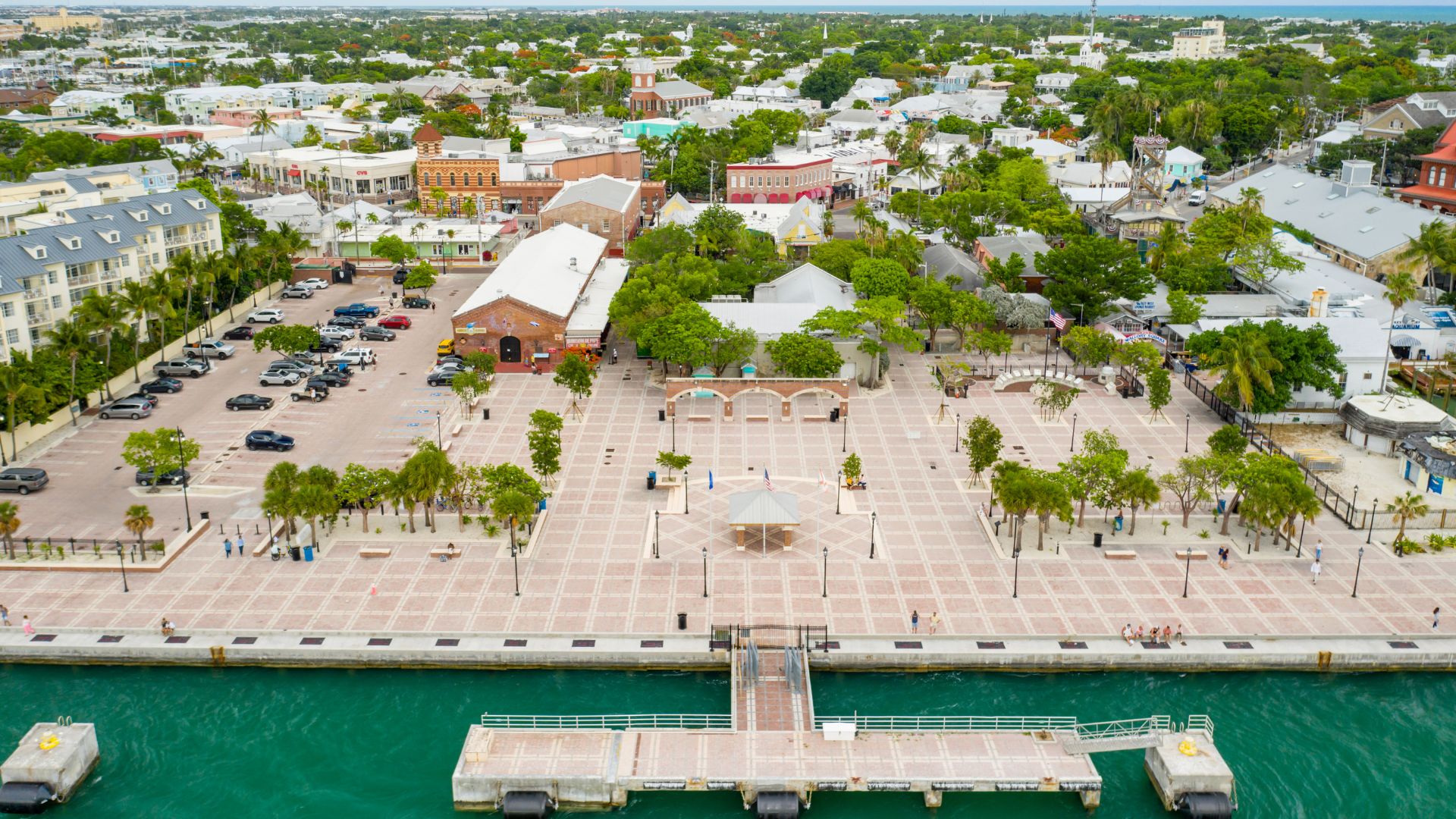 An aerial view of the paved Mallory Square plaza and pier in Key West, Florida, with several buildings and palm trees surrounding the area and turquoise water in the foreground.