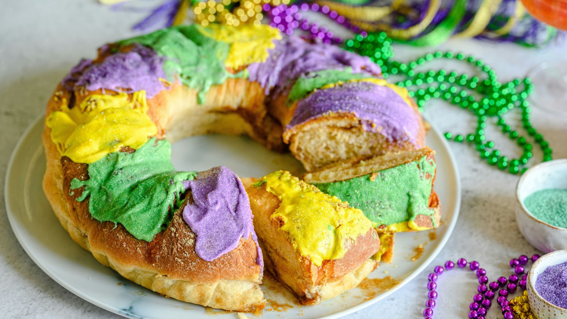 A ring-shaped Mardi Gras king cake with thick purple, green, and yellow icing and a few cut slices on a white plate, surrounded by colorful plastic beads.