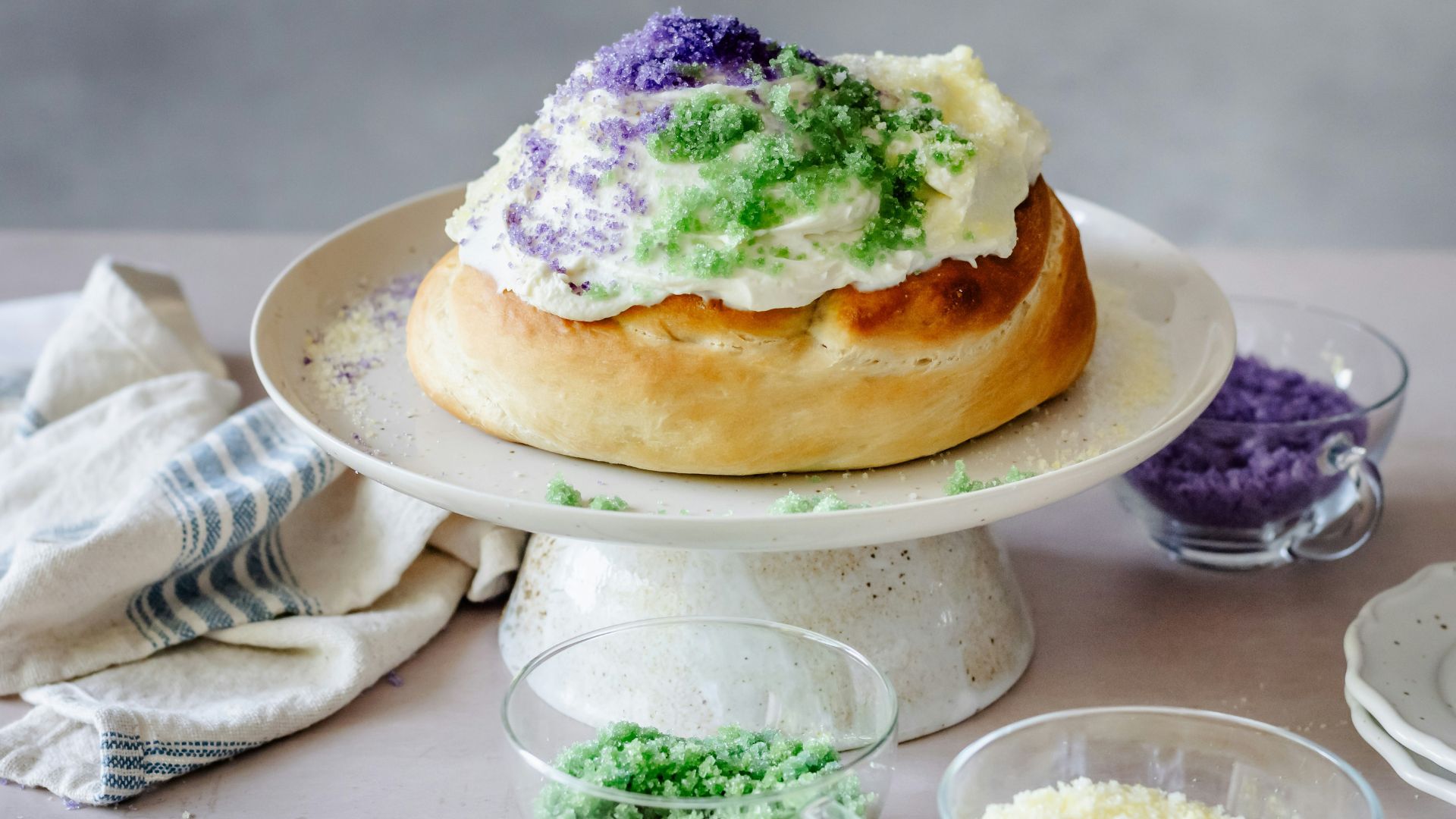 A Mardi Gras King Cake on a white cake stand, decorated with white frosting and purple, green, and yellow sprinkles. Small glass bowls containing extra purple, green, and yellow sprinkles sit on the table nearby.