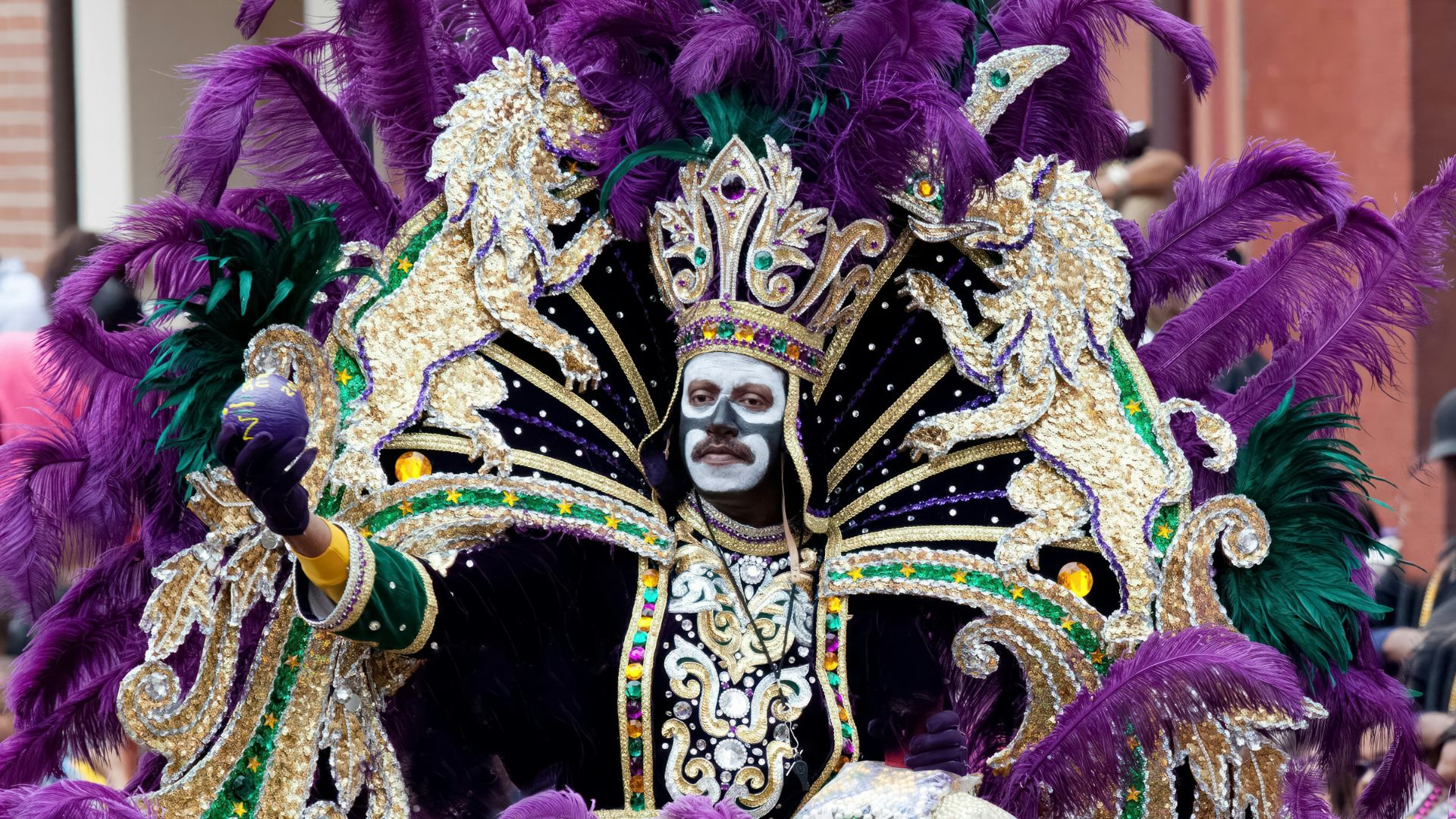 A person in an elaborate purple, green, and gold Mardi Gras costume featuring a feathered headdress, face paint, and decorative lions, is participating in a parade in New Orleans, Louisiana.