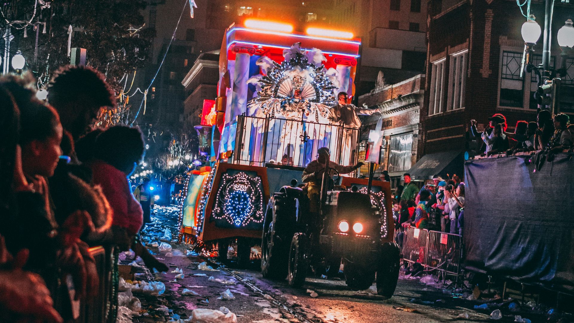 A brightly lit float is pulled by a small tractor down a wet street at night during a busy Mardi Gras parade in New Orleans. Crowds of people line the street behind temporary barricades.
