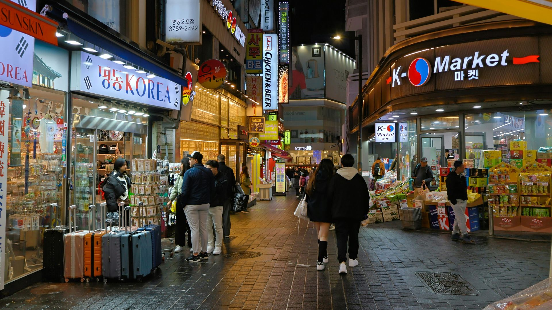 A night view of a busy commercial street, likely in Seoul, South Korea, featuring numerous brightly lit shops with Korean signage, shoppers, and people with rolling luggage.