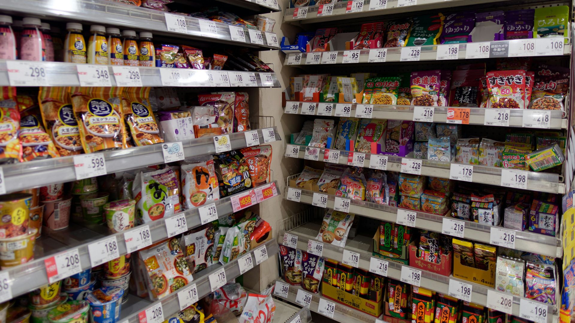 An interior view of a well-stocked Japanese convenience store or supermarket aisle, displaying numerous packaged snacks, instant foods, and drinks with Japanese labels and price tags visible on the shelves.