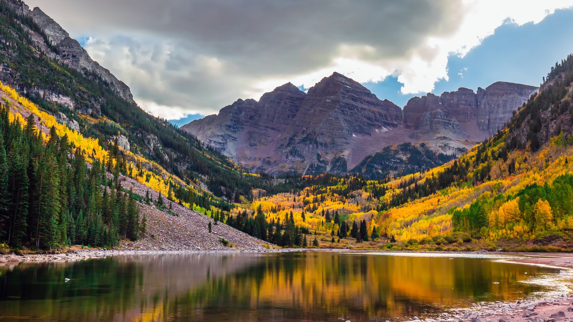 A view across the calm, reflective waters of Maroon Lake toward the iconic Maroon Bells peaks, surrounded by vibrant golden aspen trees and green pine forests under a dramatic cloudy sky during autumn in Colorado.