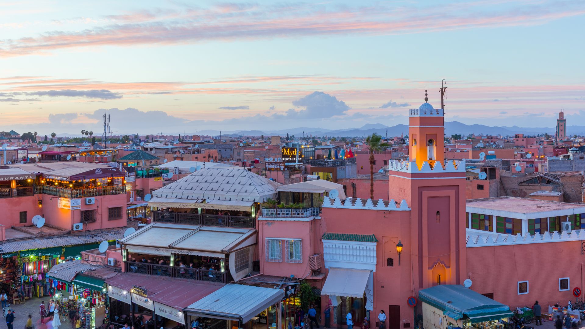 A view over the red-toned rooftops of Marrakech, Morocco, with a minaret tower prominent against the distant mountains at sunset.