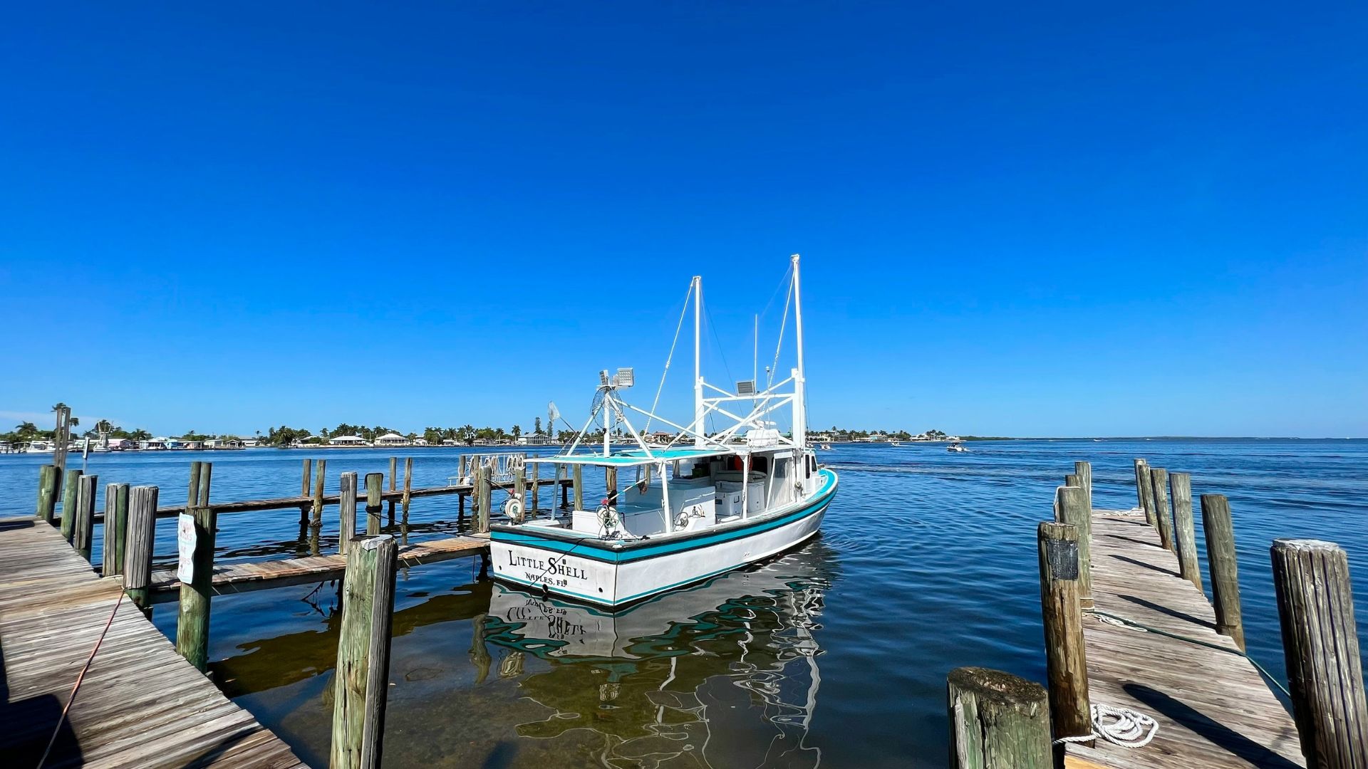 A fishing boat named 'LITTLE SHELL' is docked at a wooden pier on tranquil, shallow bay waters, with a small coastal community visible in the background under a bright blue sky.