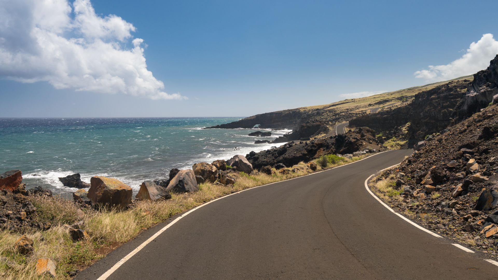 A two-lane coastal road curves along a rugged, rocky shoreline and vibrant blue ocean under a sunny sky with white clouds, likely in the Hana region of Maui, Hawaii.