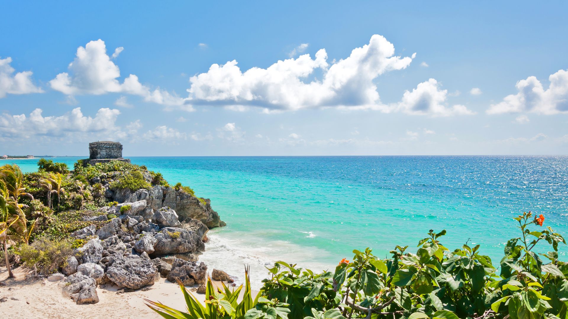 A view of the ancient Mayan ruins of Tulum, Mexico, featuring El Castillo pyramid on a cliff edge overlooking the bright blue and turquoise waters of the Caribbean Sea.