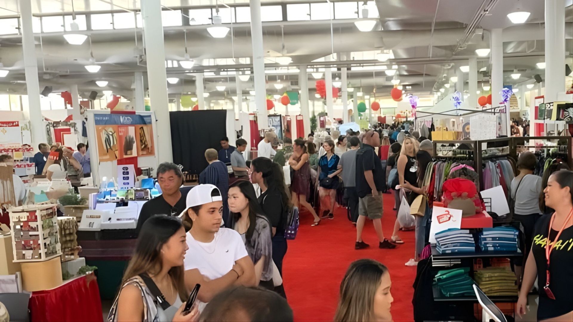 A busy indoor marketplace with many people browsing various vendor booths and merchandise under bright overhead lights.