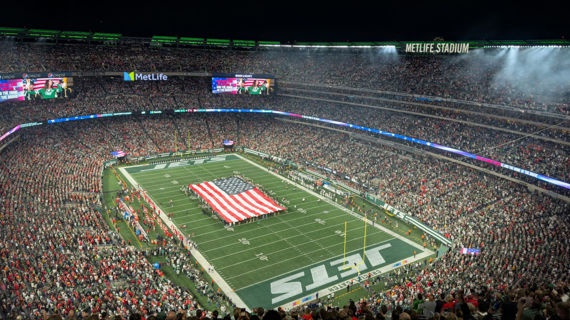 A crowded nighttime view of the MetLife Stadium football field, with an American flag displayed on the turf and the New York Jets logo visible in the end zone.