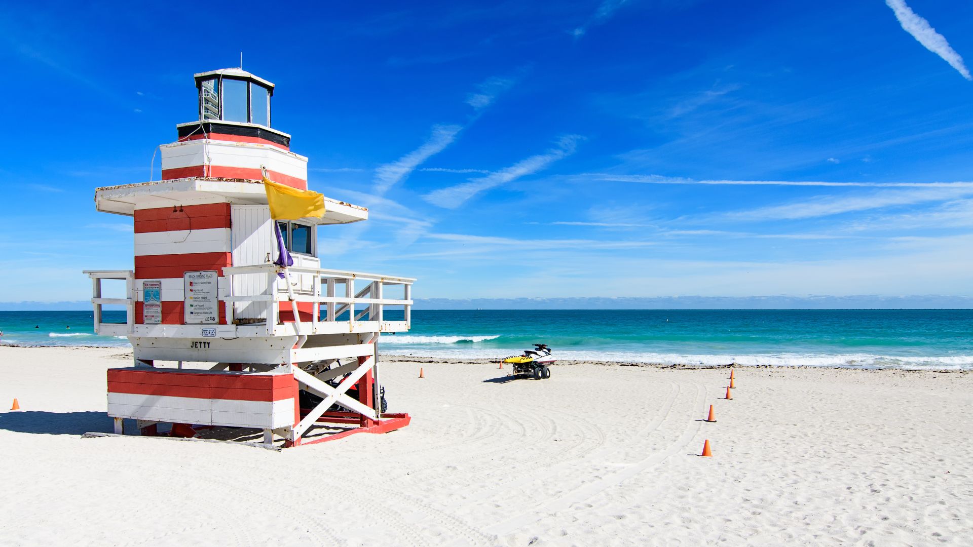 A red and white striped lifeguard stand on a white sand beach with turquoise water and a blue sky.