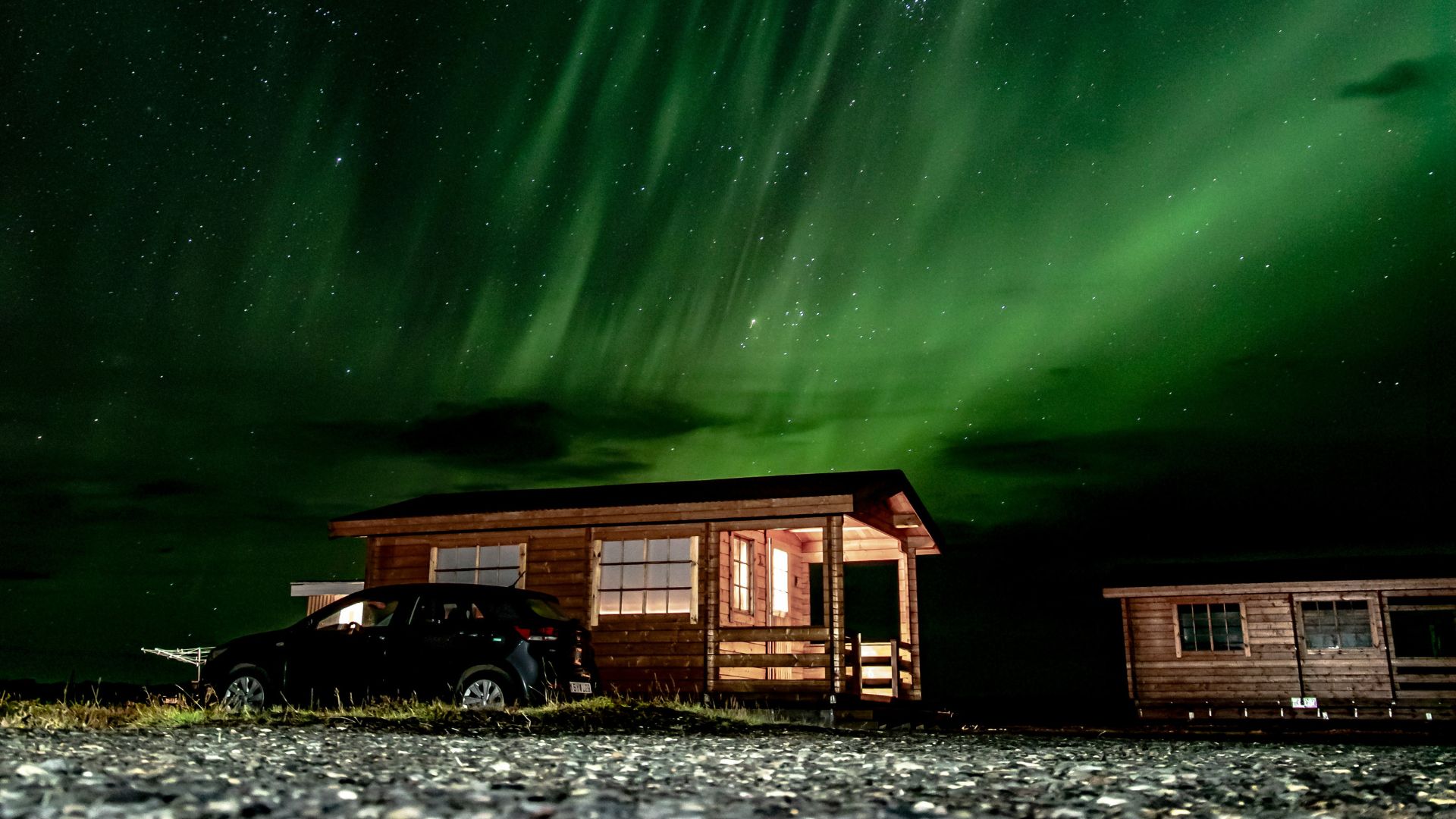 A black SUV is parked in front of two wooden cabins under a vibrant green and purple aurora borealis in Iceland.