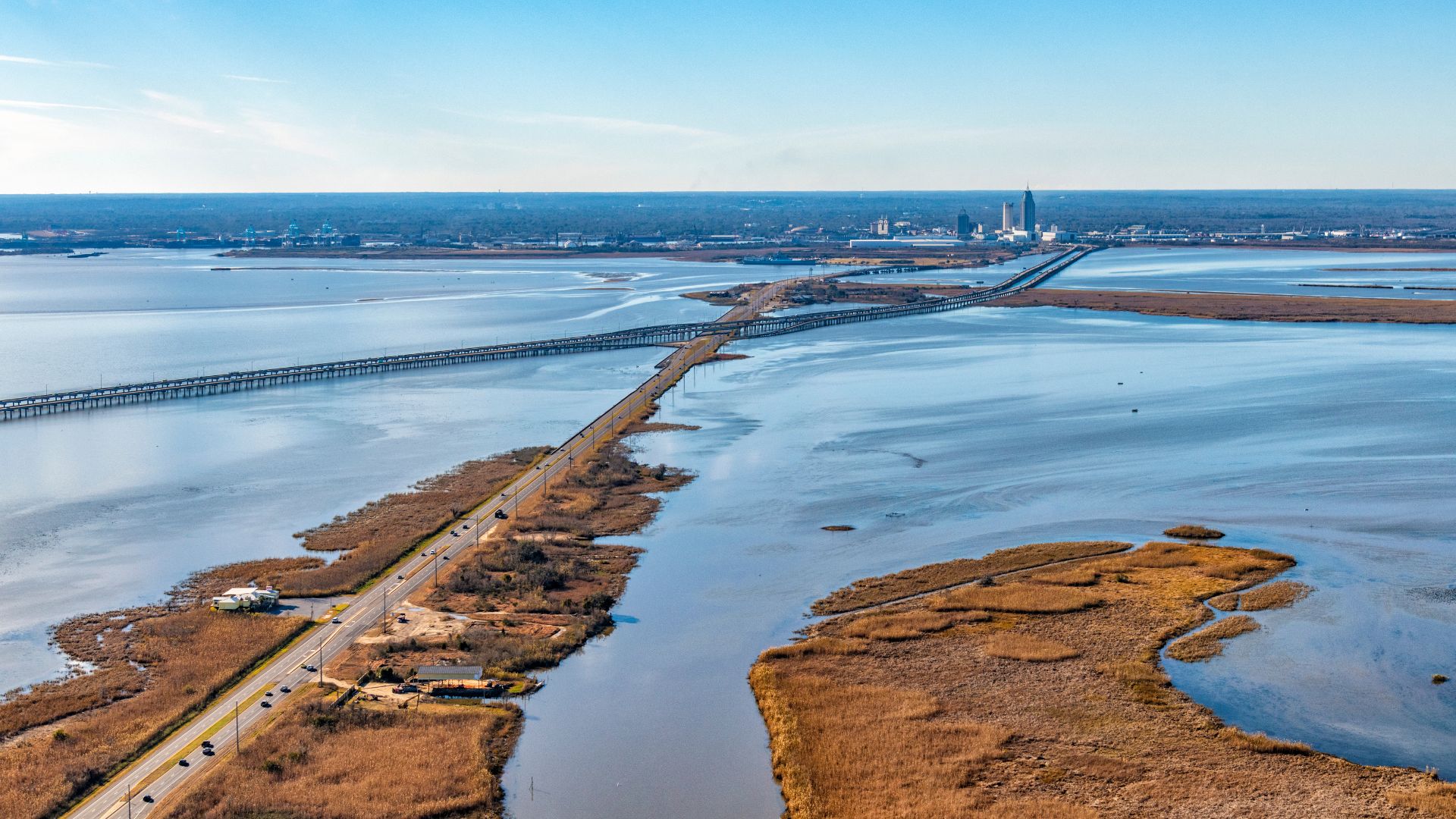 An aerial view of a long highway causeway and a parallel bridge spanning a wide bay, with marshy islands covered in brown and green grass in the foreground and a distant city skyline under a clear blue sky.