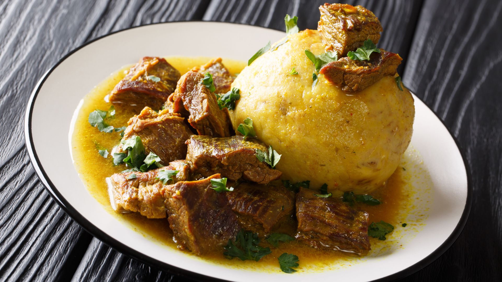 Mofongo (mashed fried plantains with pork cracklings) is served alongside a savory beef or pork stew in a white plate on a dark wooden table.