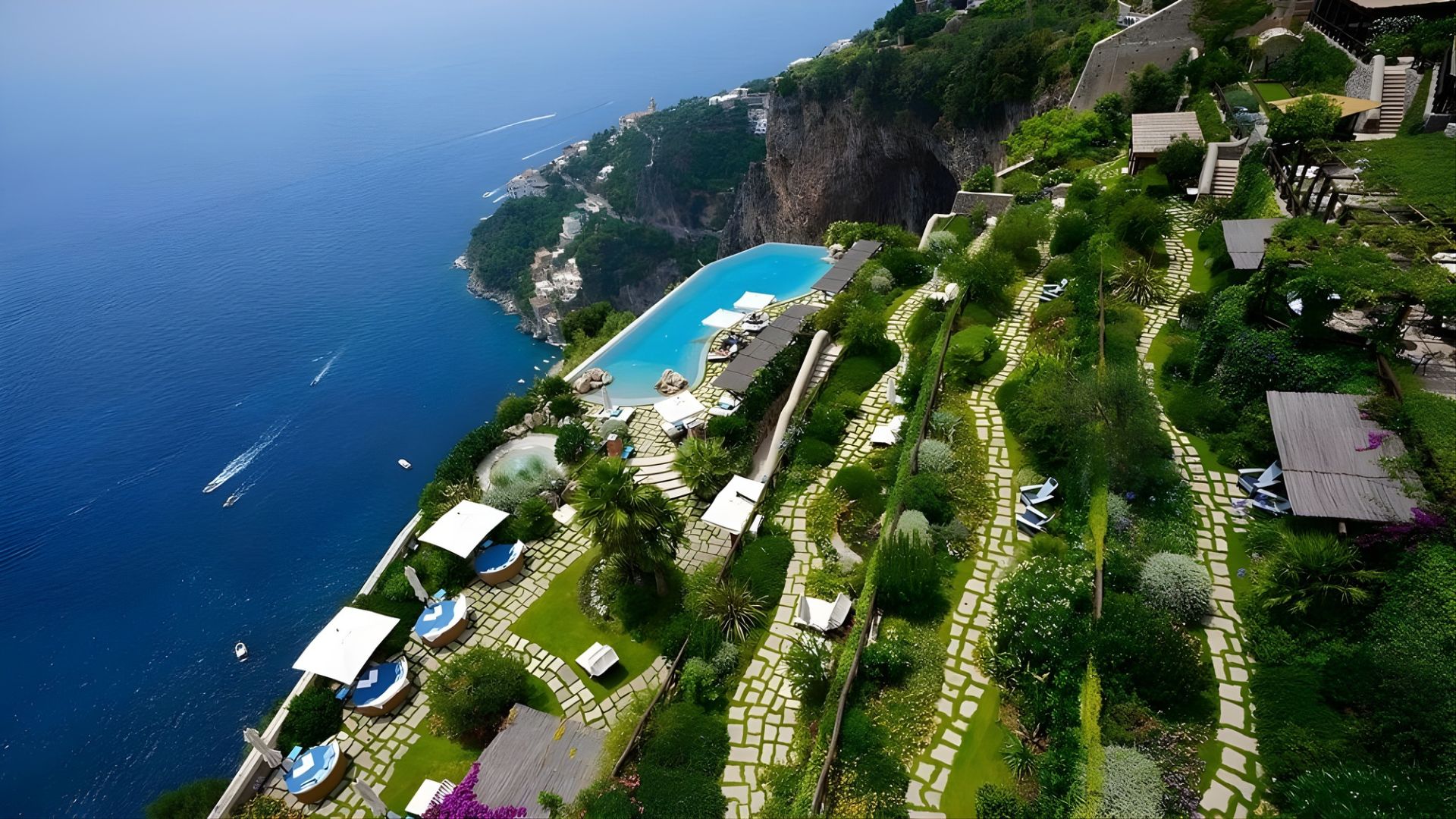 An aerial view of a luxury hotel, Monastero Santa Rosa, built into a lush green cliffside, featuring a large infinity pool and terraced gardens overlooking the deep blue sea of the Amalfi Coast.