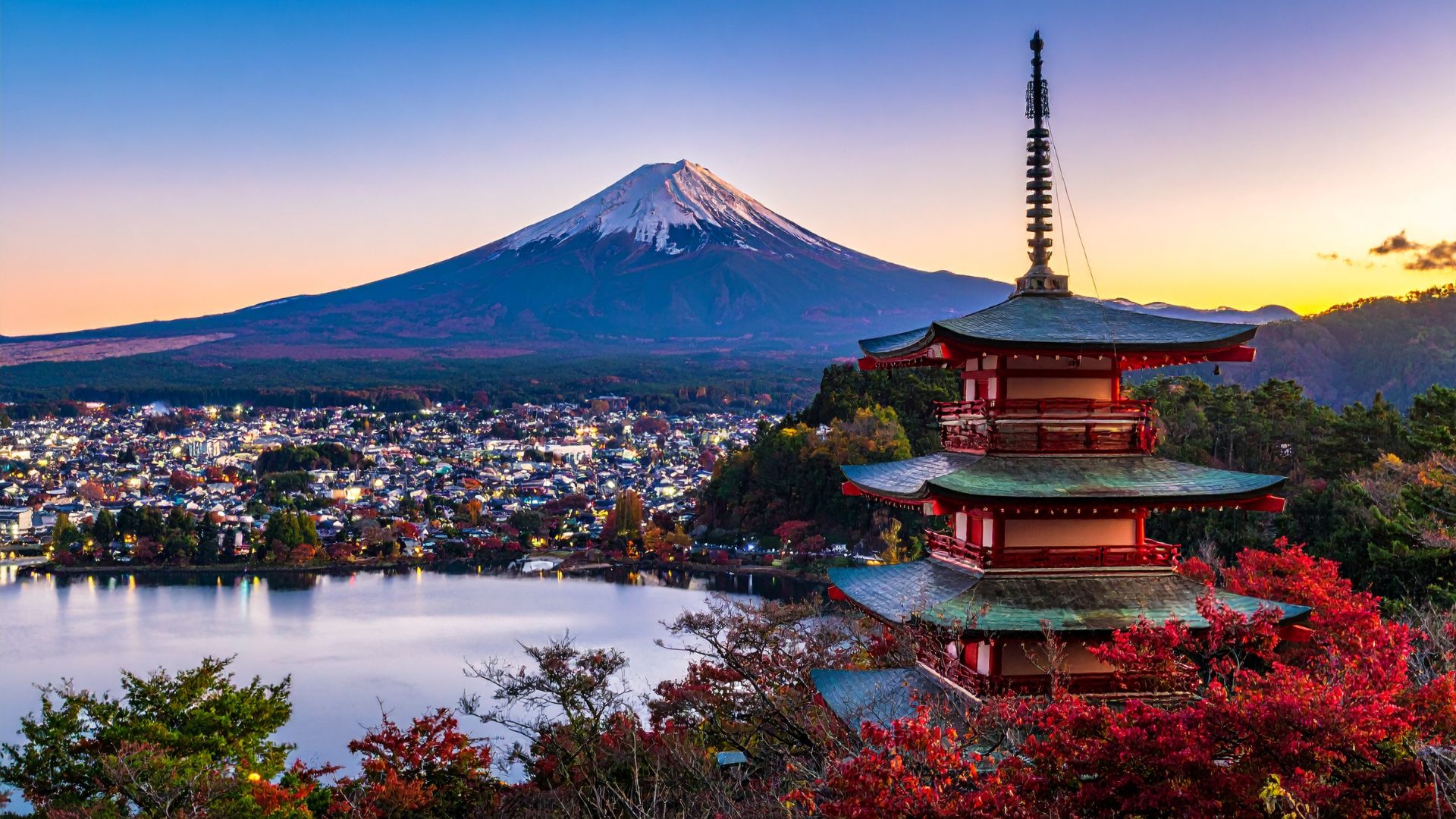 A view of the Chūreitō Pagoda with autumn foliage in the foreground and the snow-capped Mount Fuji in the background under a blue sky at sunset.