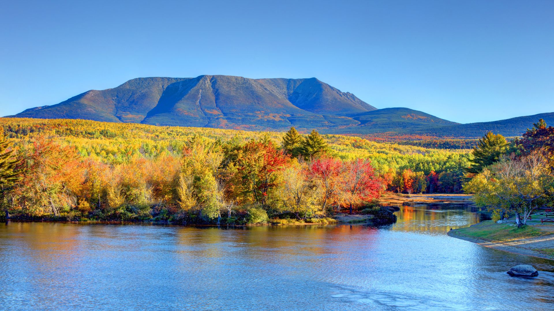 A wide, scenic view of the massive Mount Katahdin rising in the background, with calm blue water reflecting the mountain and a shoreline of dense forest with vibrant yellow, orange, and red fall foliage in the foreground.