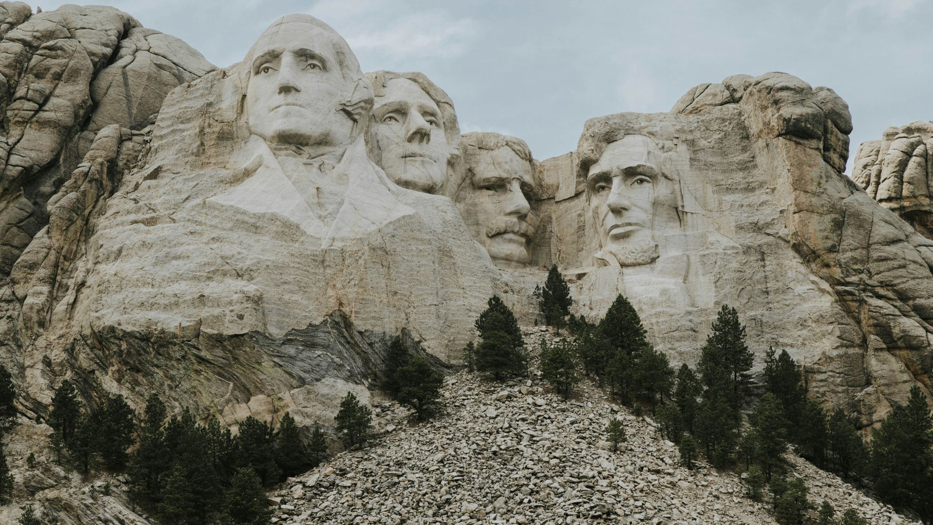A close view of the carved faces of four U.S. presidents on the granite cliff of Mount Rushmore, with a rocky slope and green evergreen trees in the foreground.