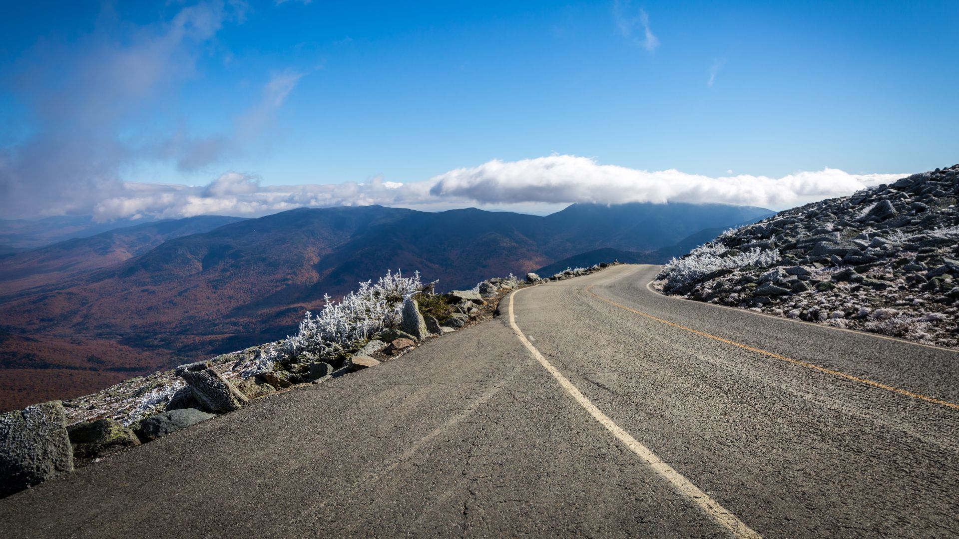 A two-lane mountain highway with no guardrails curves along a rugged, rocky cliff edge with white frost, overlooking an autumnal valley and distant mountains under a blue sky.