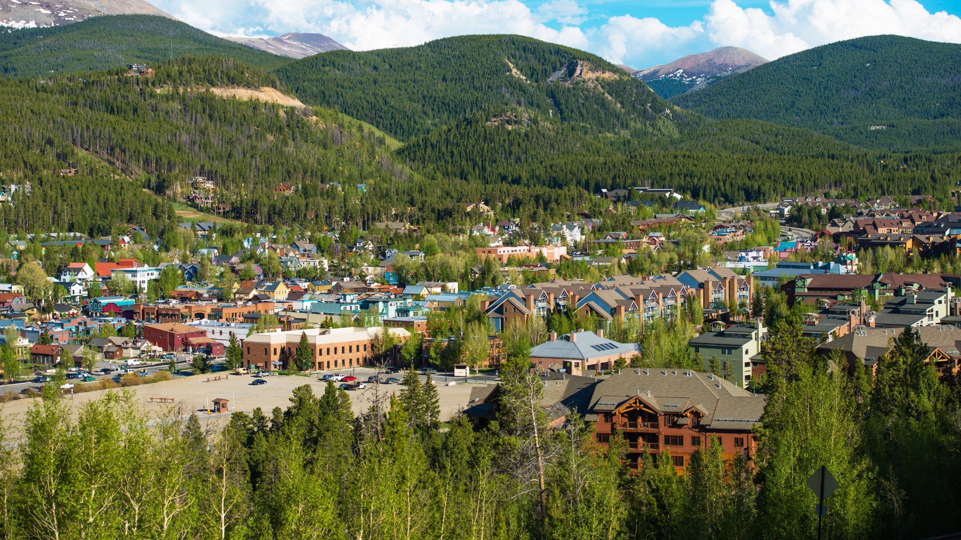 An aerial, summer view of the mountain town of Breckenridge, Colorado, surrounded by green, tree-covered mountains under a bright blue sky.