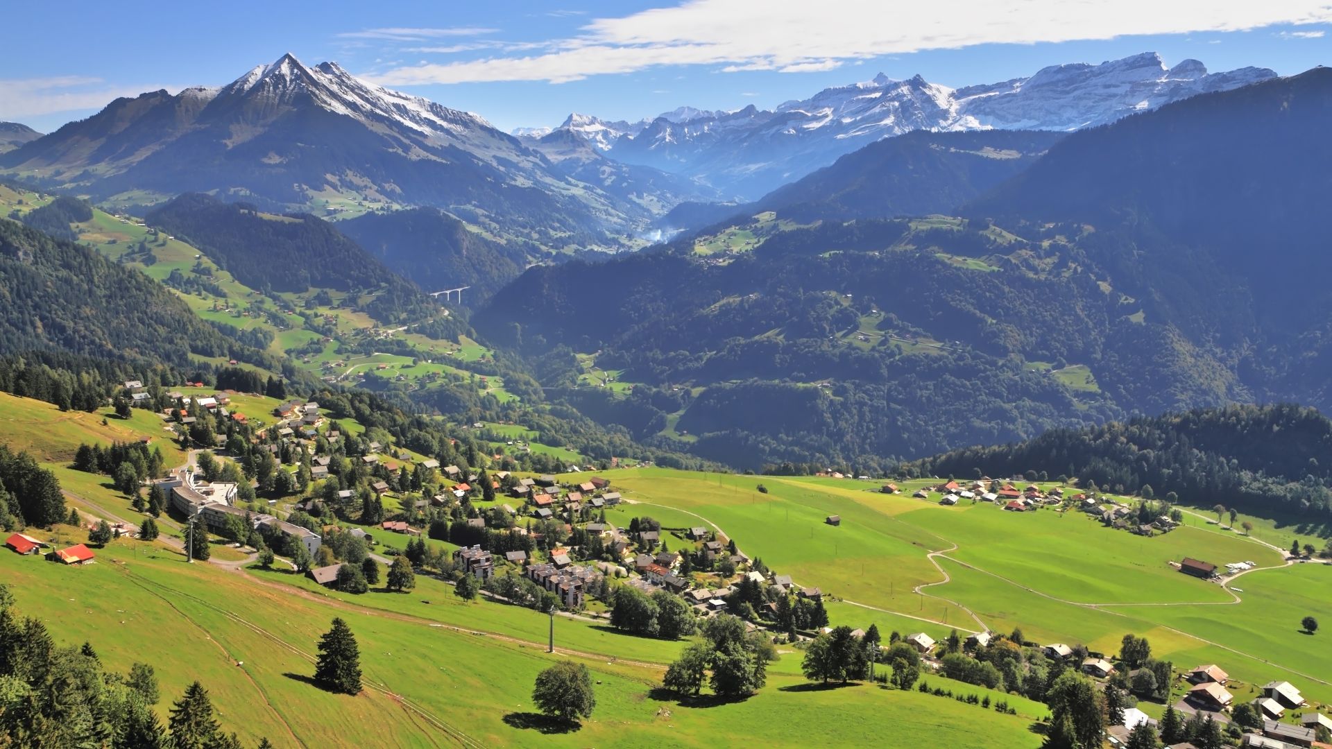 An elevated, wide view of the Swiss mountain village of Leysin nestled on a green hillside, surrounded by steep, tree-covered slopes and tall, snow-capped alpine peaks in the distance.