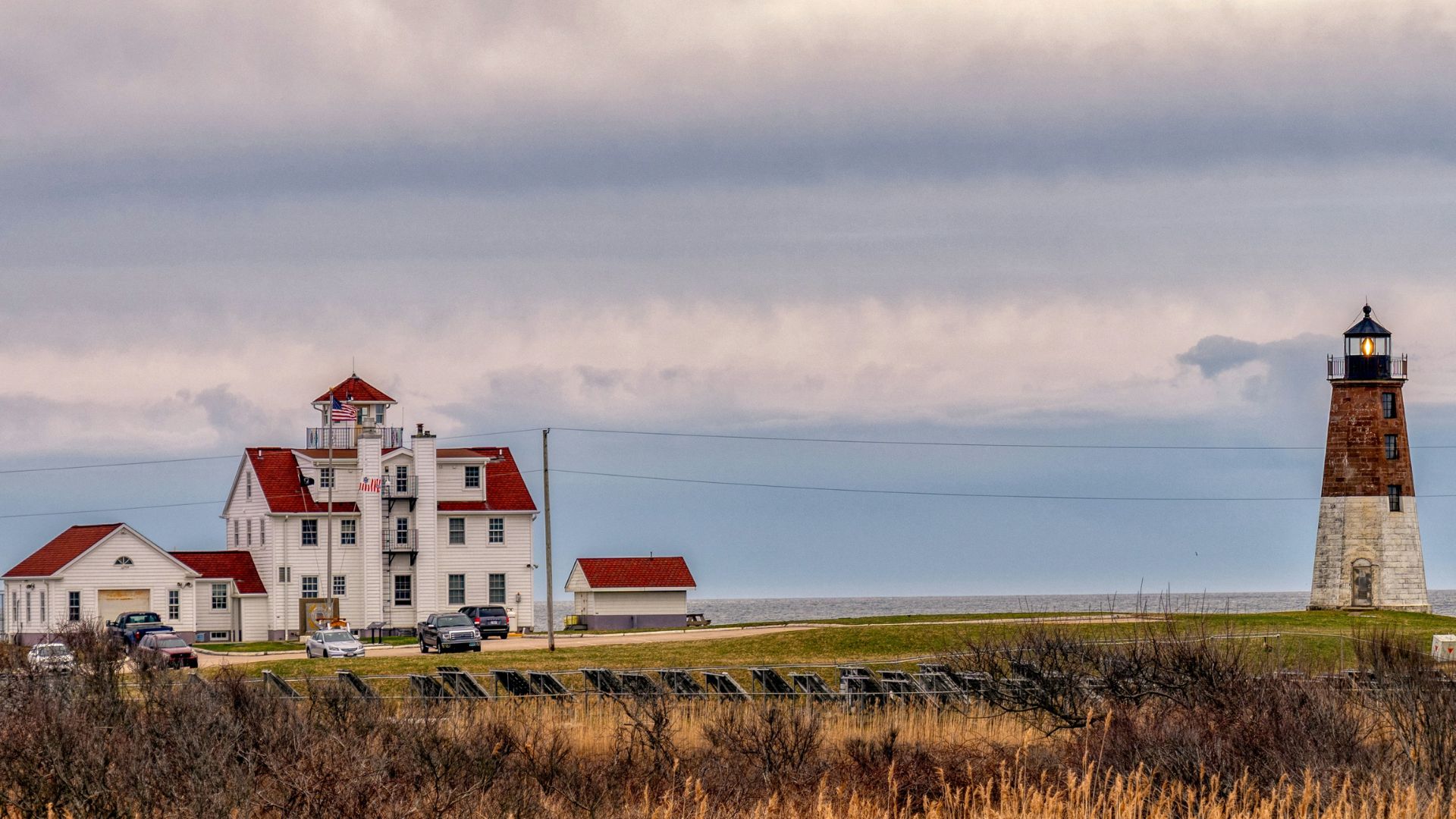 A historic brown and white octagonal lighthouse and a large white building on a grassy point of land bordering the ocean under a cloudy sky.