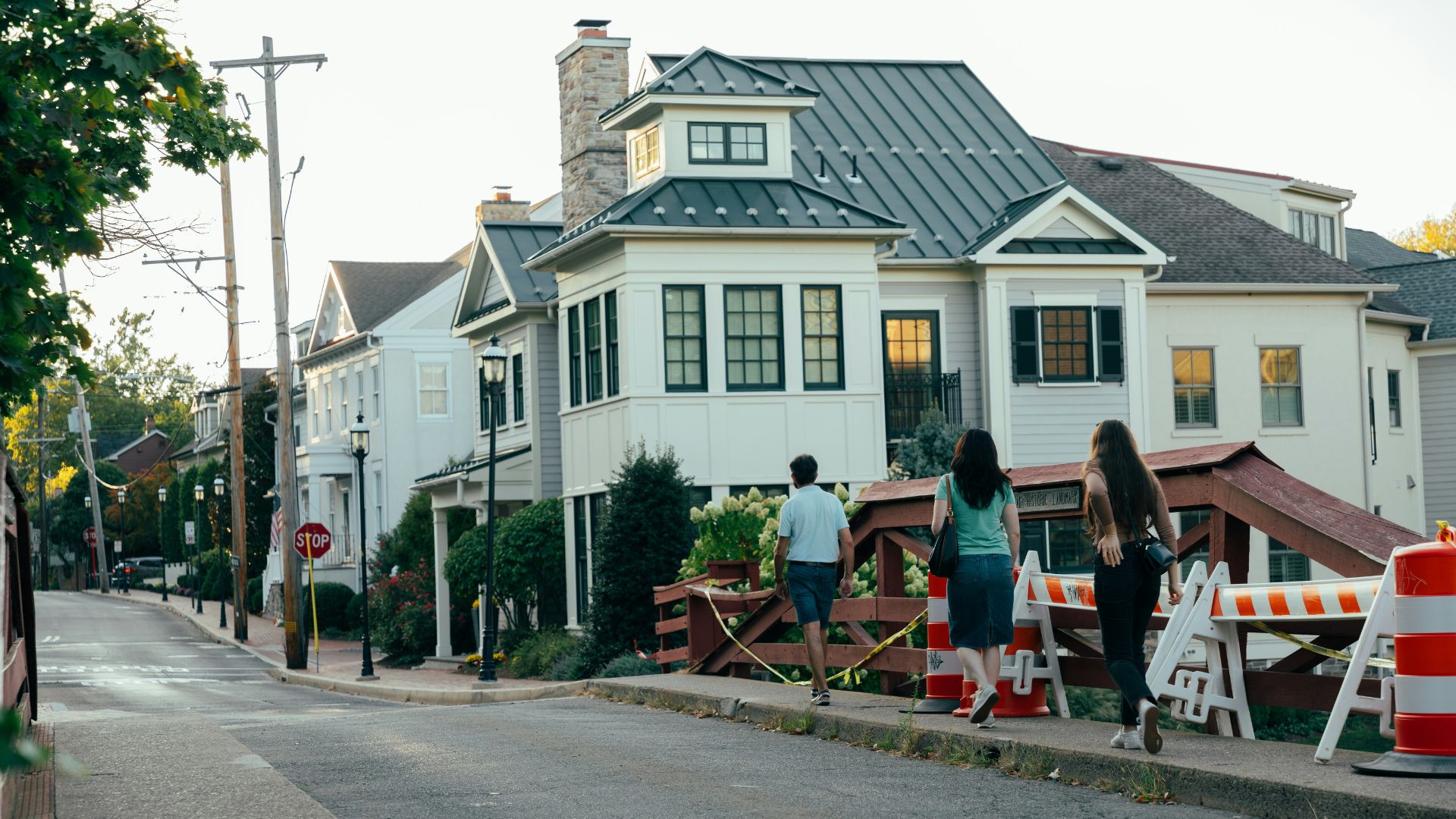A view of a quiet American residential street with several two-story houses featuring white siding and dark metal roofs. Three people are walking away from the camera on the sidewalk, passing construction barriers near the curb as the sun sets.