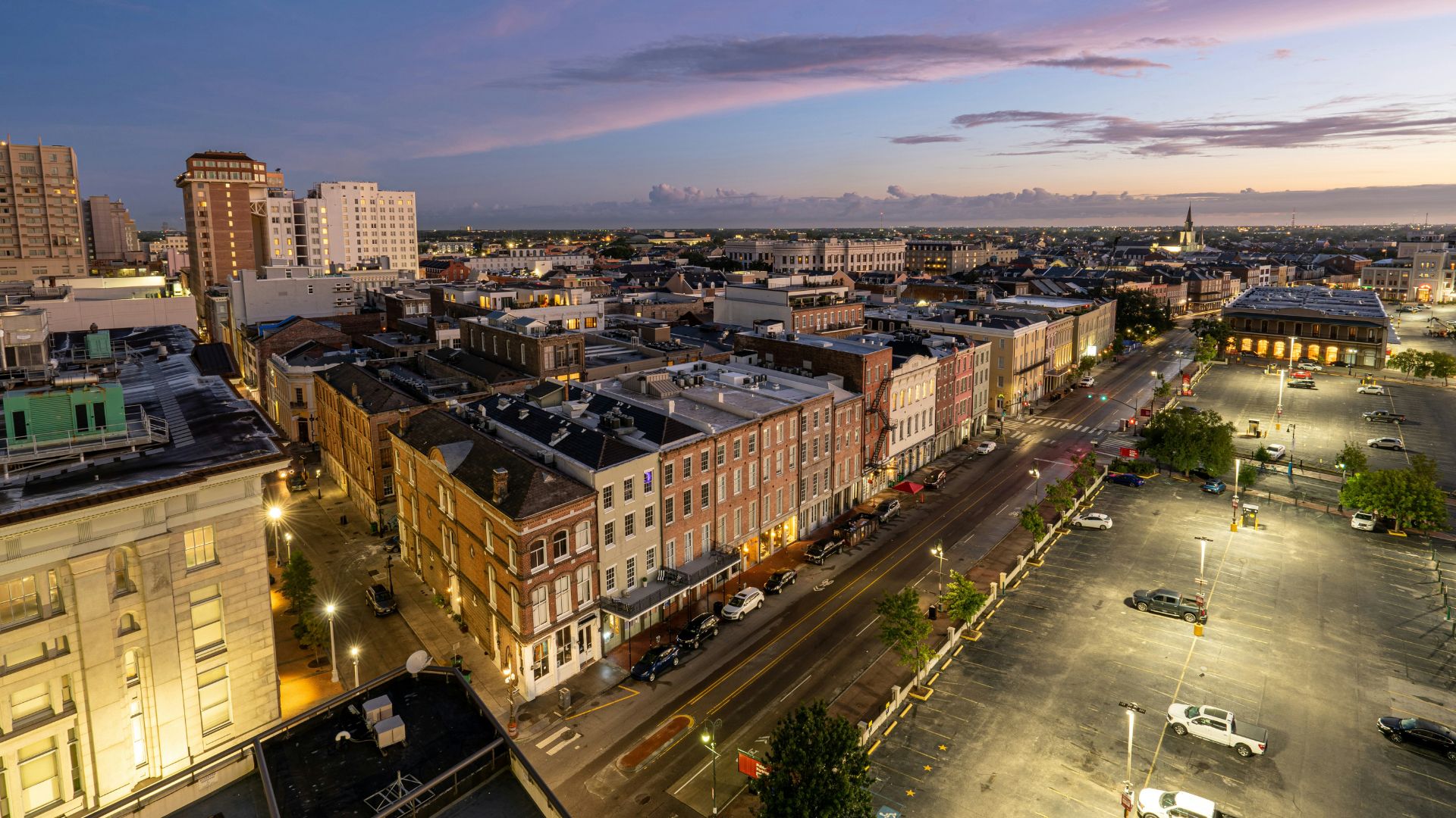 An elevated view of the historic French Quarter in New Orleans at dusk, showing rows of colorful traditional buildings with lighted windows, a street and large parking lot illuminated by streetlights, and taller modern buildings in the distance against a pink and blue twilight sky.