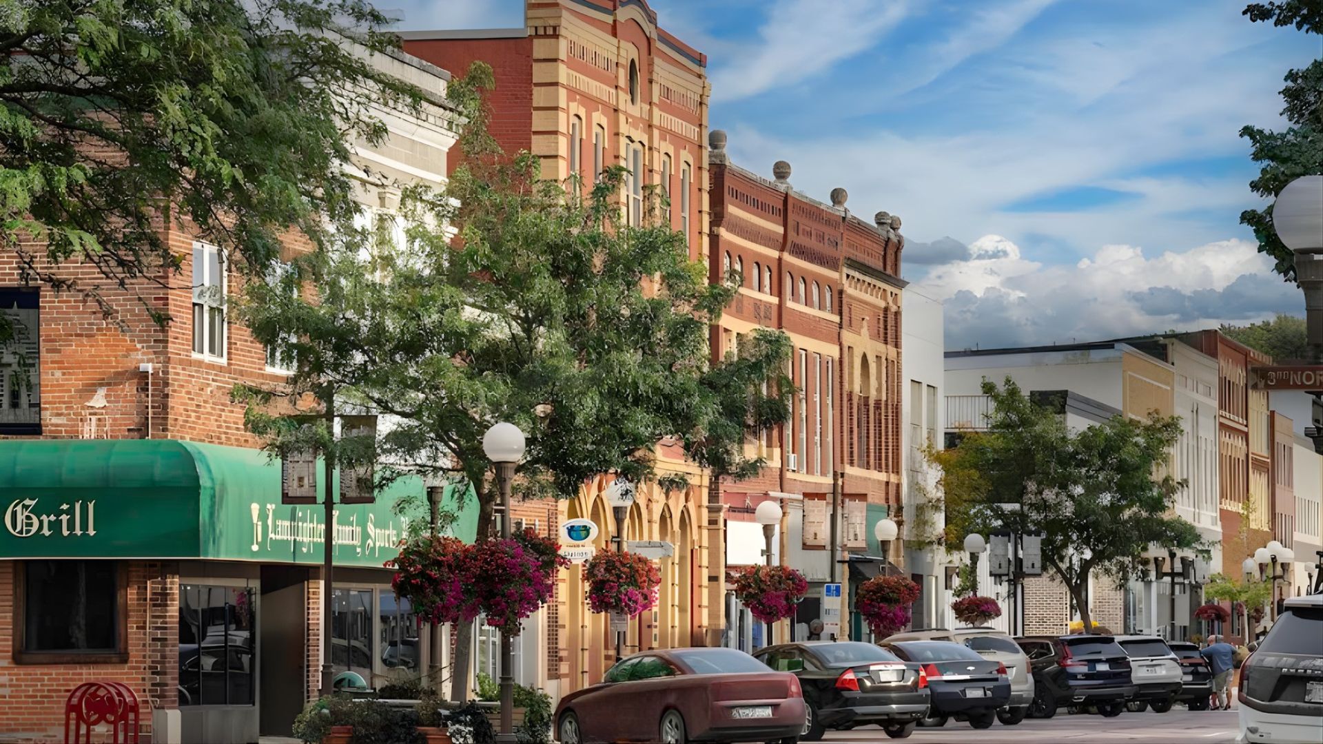 A daytime street scene in the historic downtown of New Ulm, Minnesota, showing several Victorian-era brick buildings with hanging flower baskets along a street lined with parked cars and green trees under a partly cloudy sky.