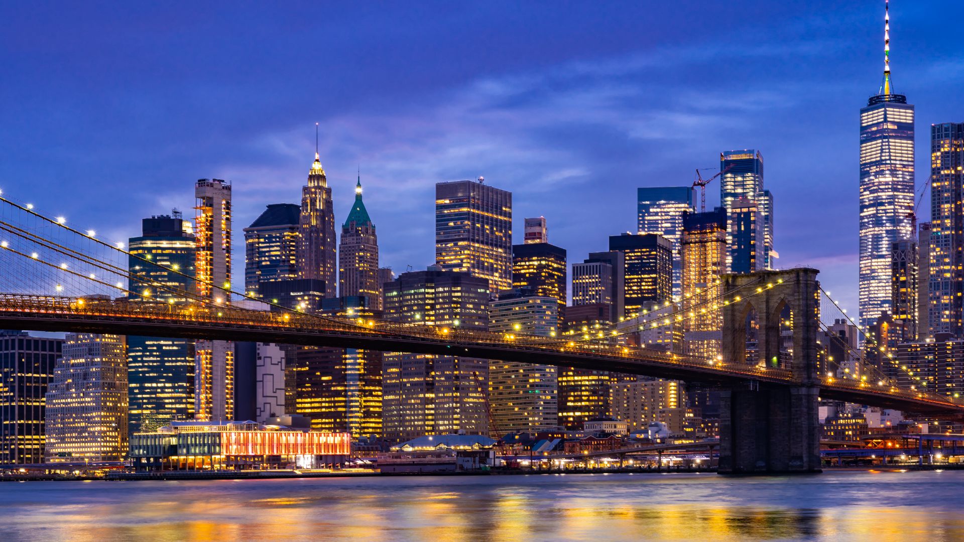 A nighttime view across the water toward the illuminated Brooklyn Bridge and the towering skyscrapers of the Manhattan skyline.
