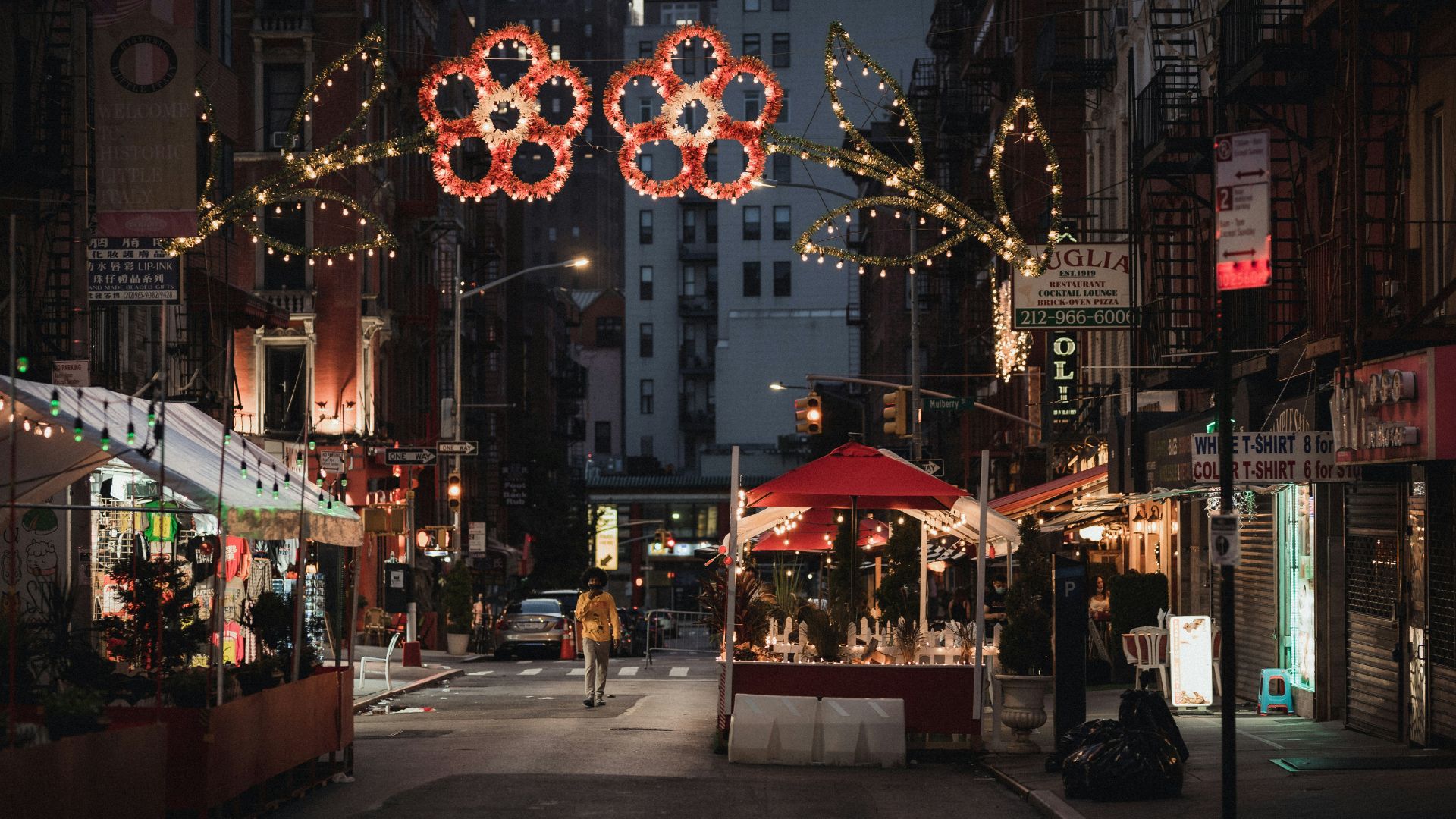 A nighttime street view of Mulberry Street in Little Italy, New York City, adorned with festive lights and outdoor dining structures.