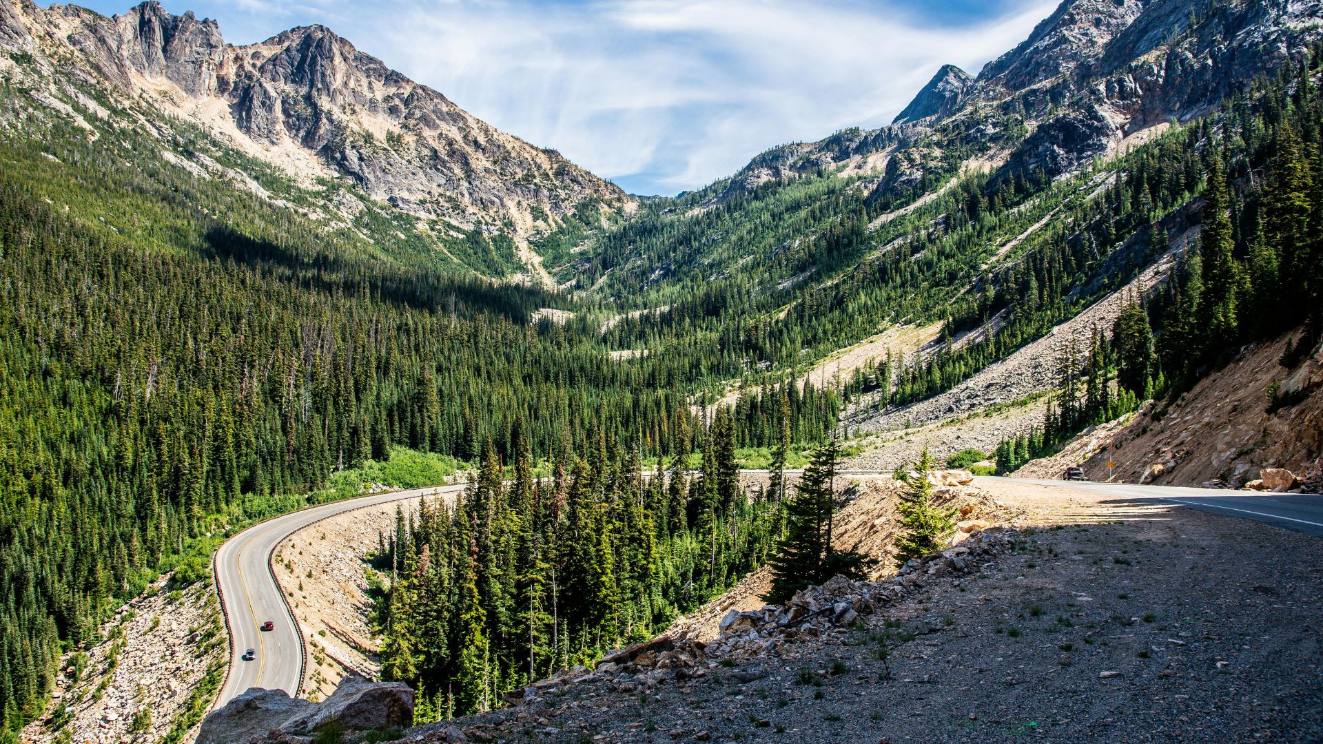An elevated view of a winding two-lane mountain highway cutting through dense evergreen forests and rugged, rocky mountains under a partly cloudy sky.