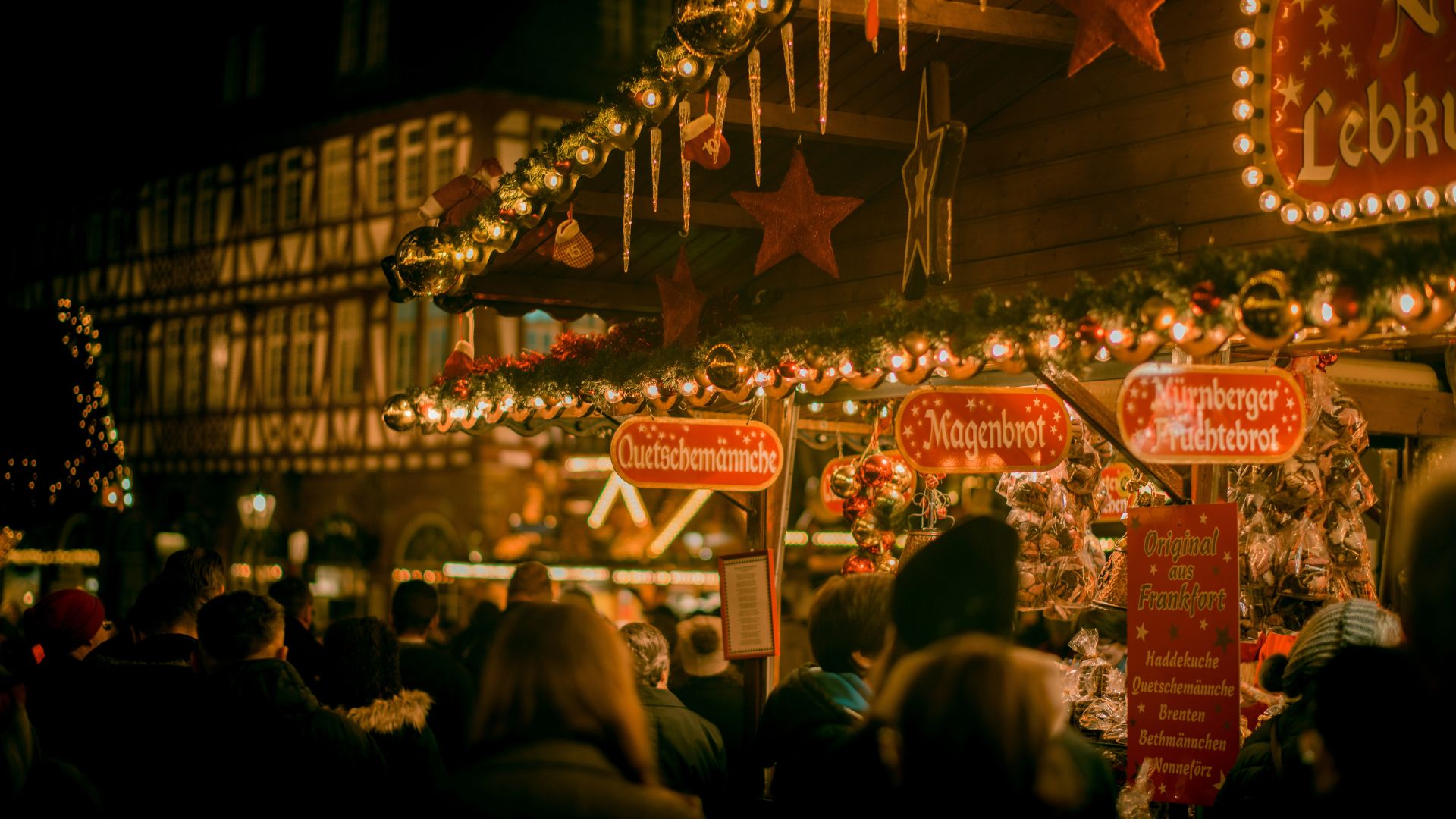 A crowd of people shops at brightly lit wooden stalls selling "Nürnberger Fruchtebrot," "Magenbrot," and "Quetschemännche" at a German Christmas market at night.