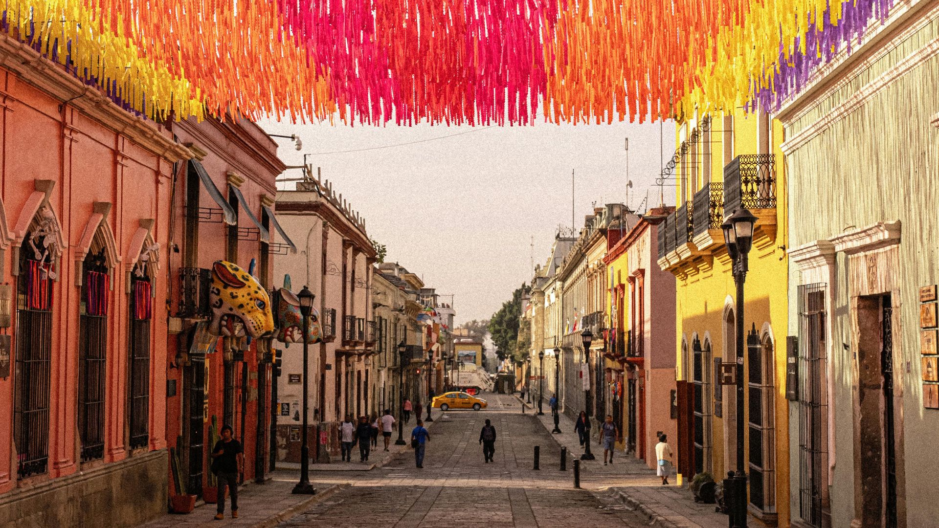 A person walking down a vibrant, colorful cobblestone street in Oaxaca, Mexico, lined with colonial-style buildings under a clear blue sky.