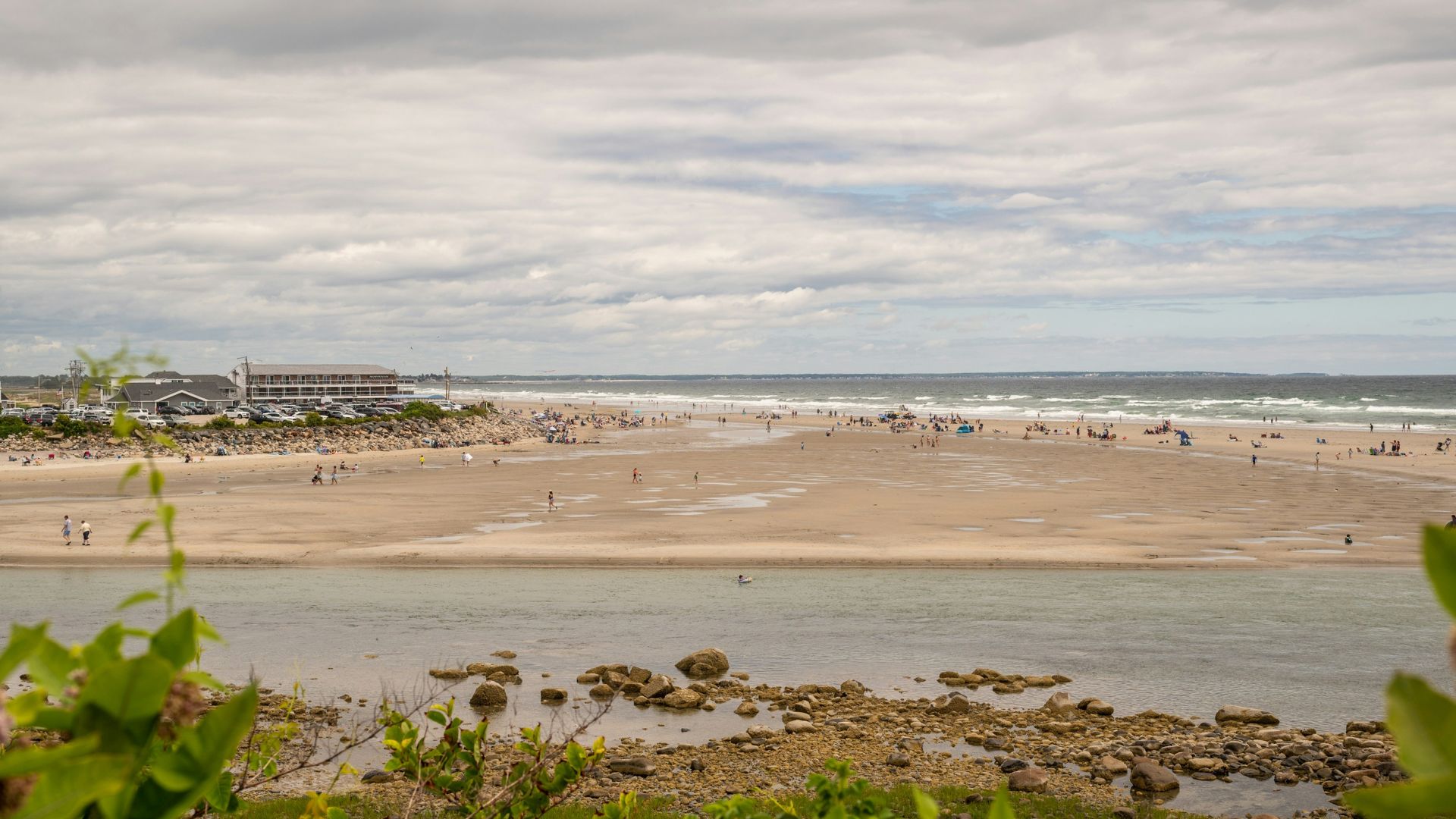 An aerial view of a wide, sandy beach at low tide with a tidal river running through it, several beachfront hotel buildings in the distance on the left, and lush green vegetation in the foreground.