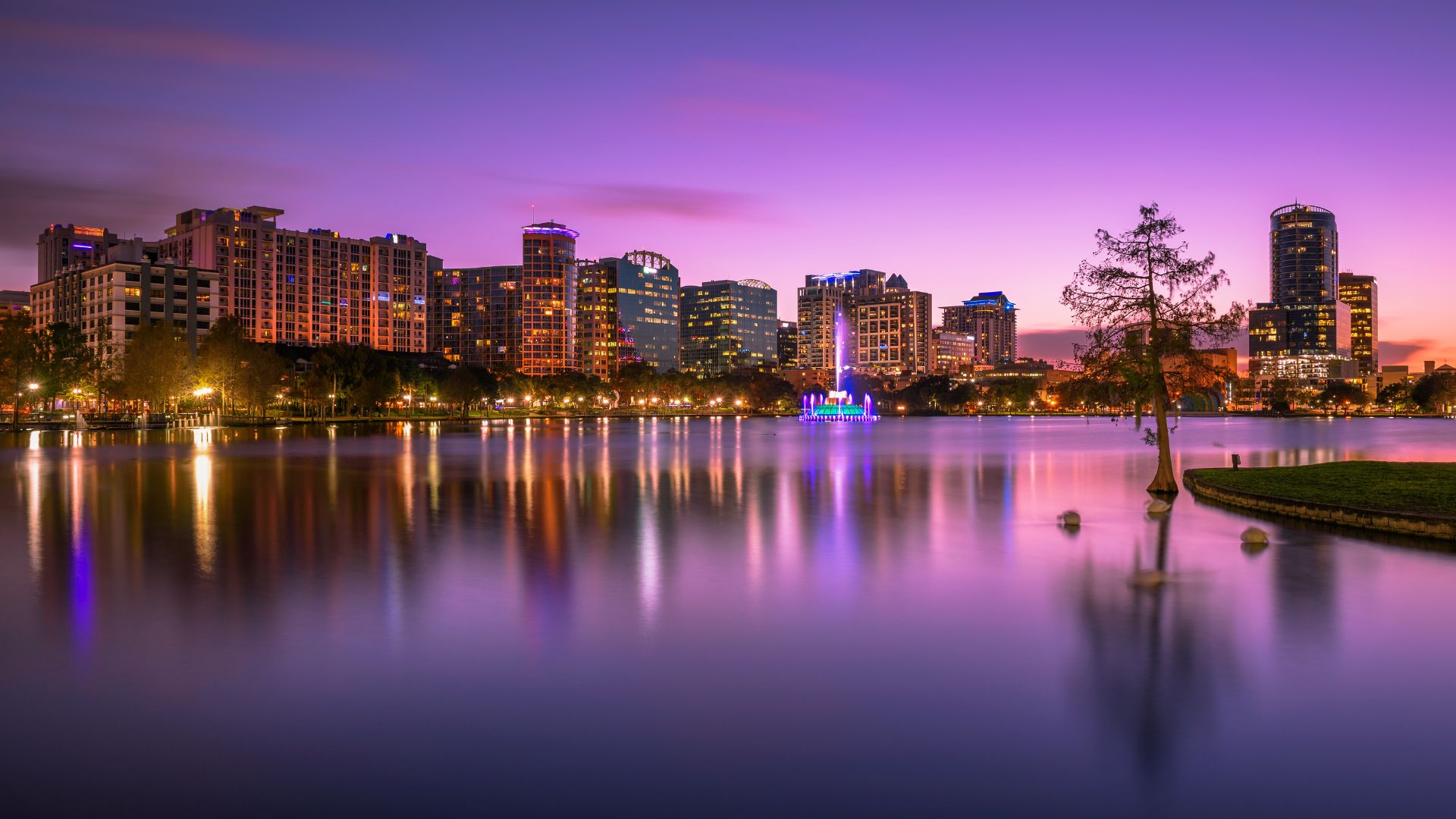 A view across the calm waters of Lake Eola reflecting the illuminated downtown Orlando skyline and a colorful central fountain at dusk.