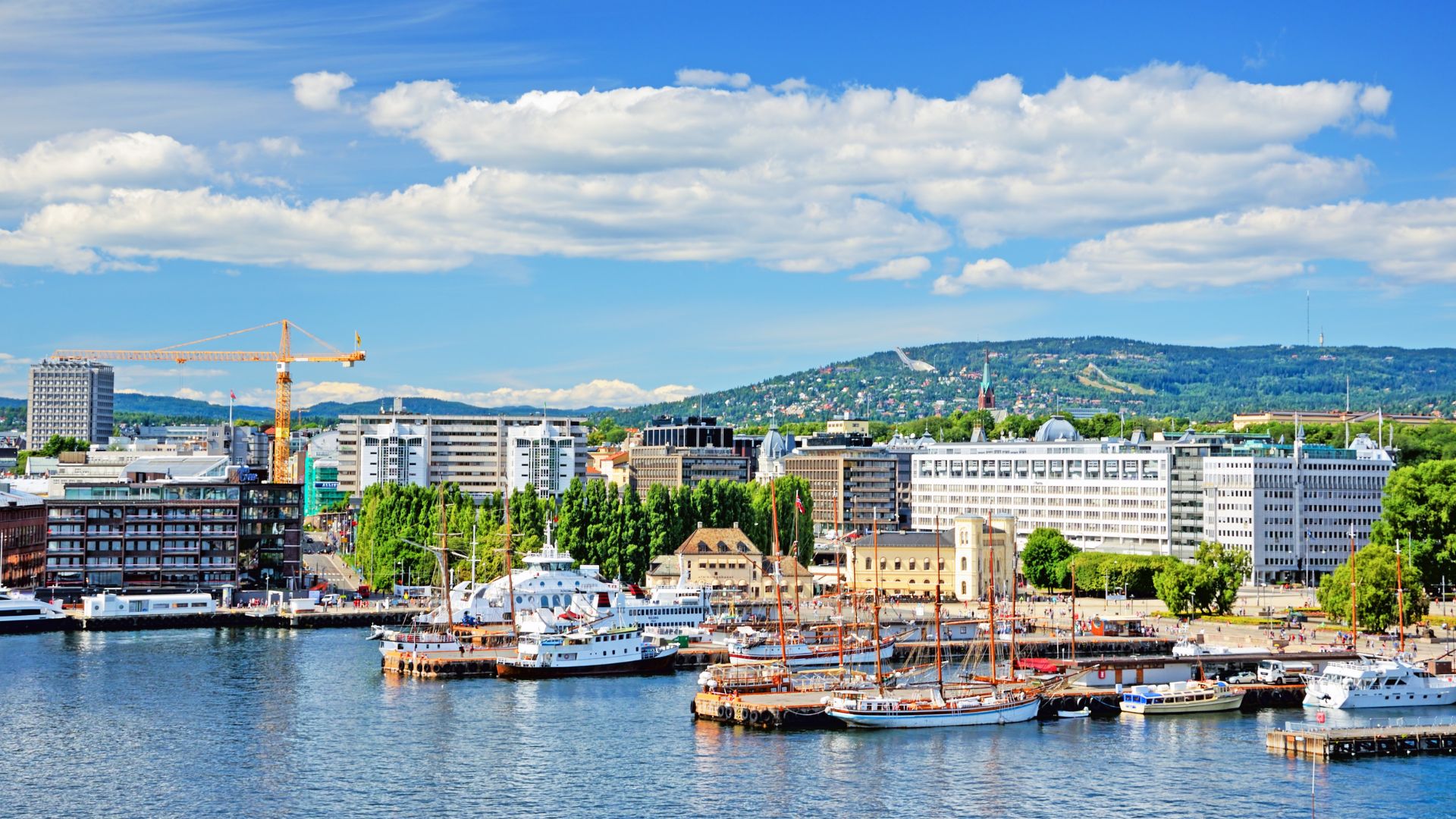 A bright, daytime photograph of the Oslo, Norway harbor featuring a blend of modern white and older brick buildings, several docked boats including a historic sailboat, a large orange construction crane, and tree-covered mountains in the background.