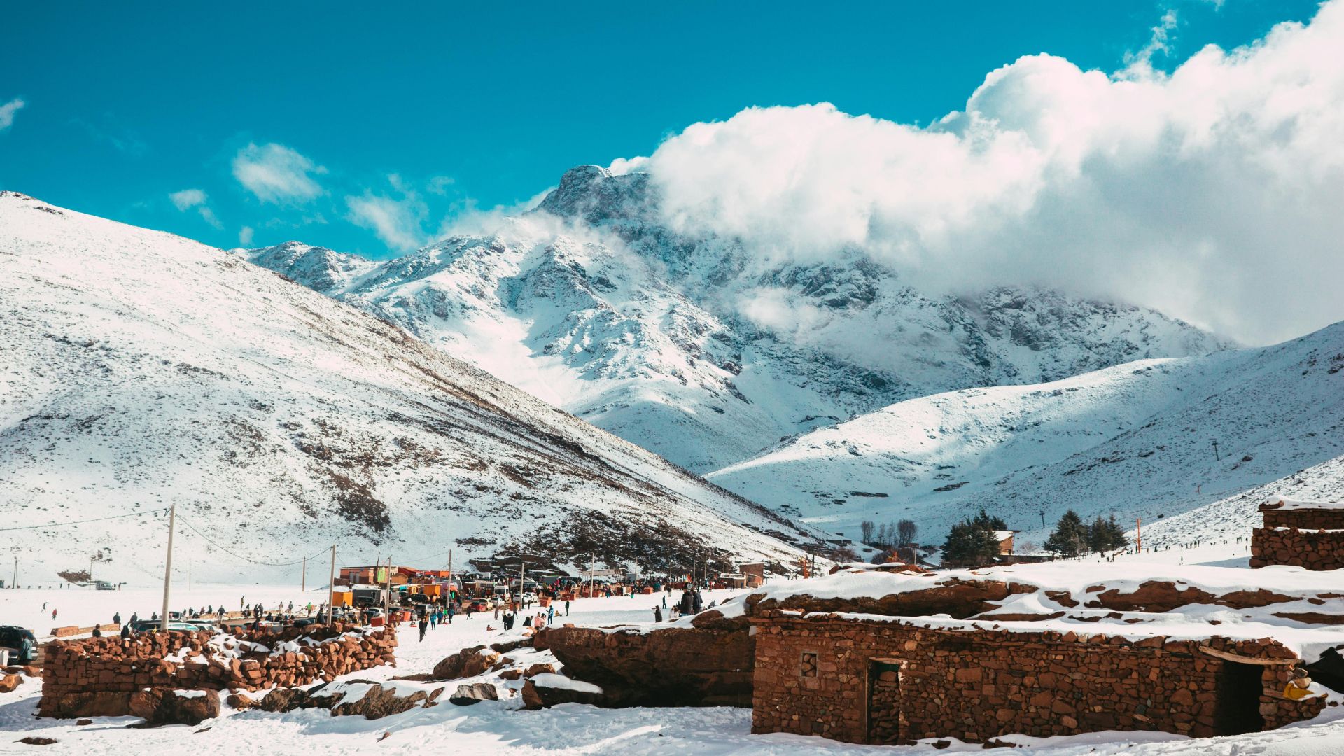 A small village with stone buildings at the base of snow-capped mountains under a bright blue sky with white clouds in the High Atlas region of Morocco.