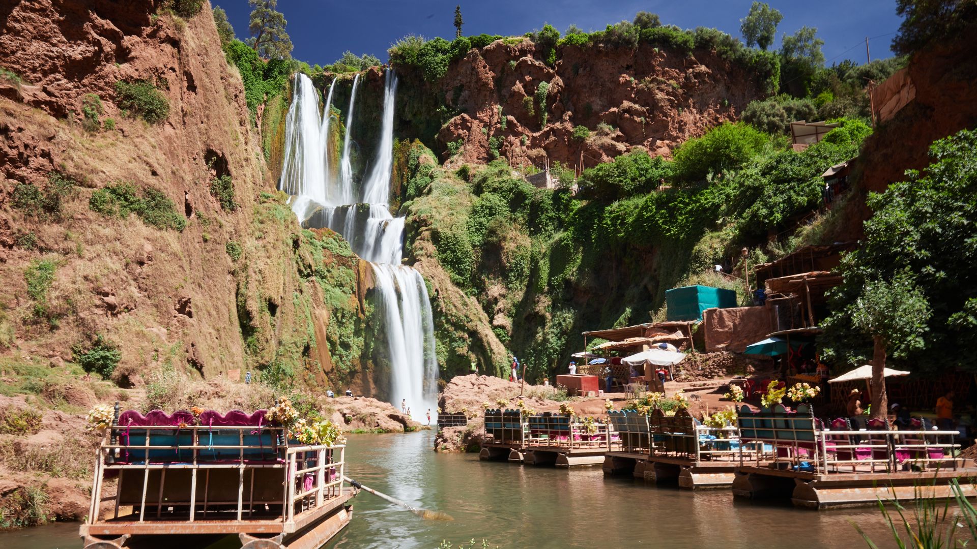 A scenic view of multi-tiered waterfalls cascading down red-rock cliffs covered in lush green vegetation, with several small, colorful tour boats docked in the pool at the base.
