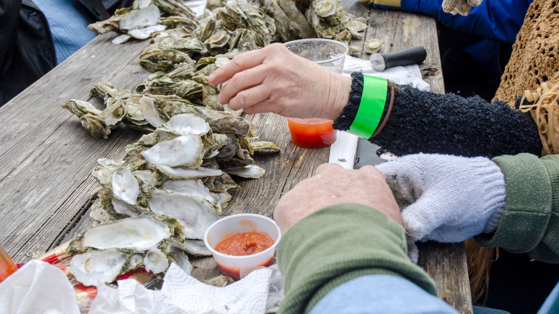 People gathered around a rustic wooden table piled high with fresh oysters, using a knife to shuck the shells, with small cups of cocktail sauce and a bright green wristband visible on one person's wrist.