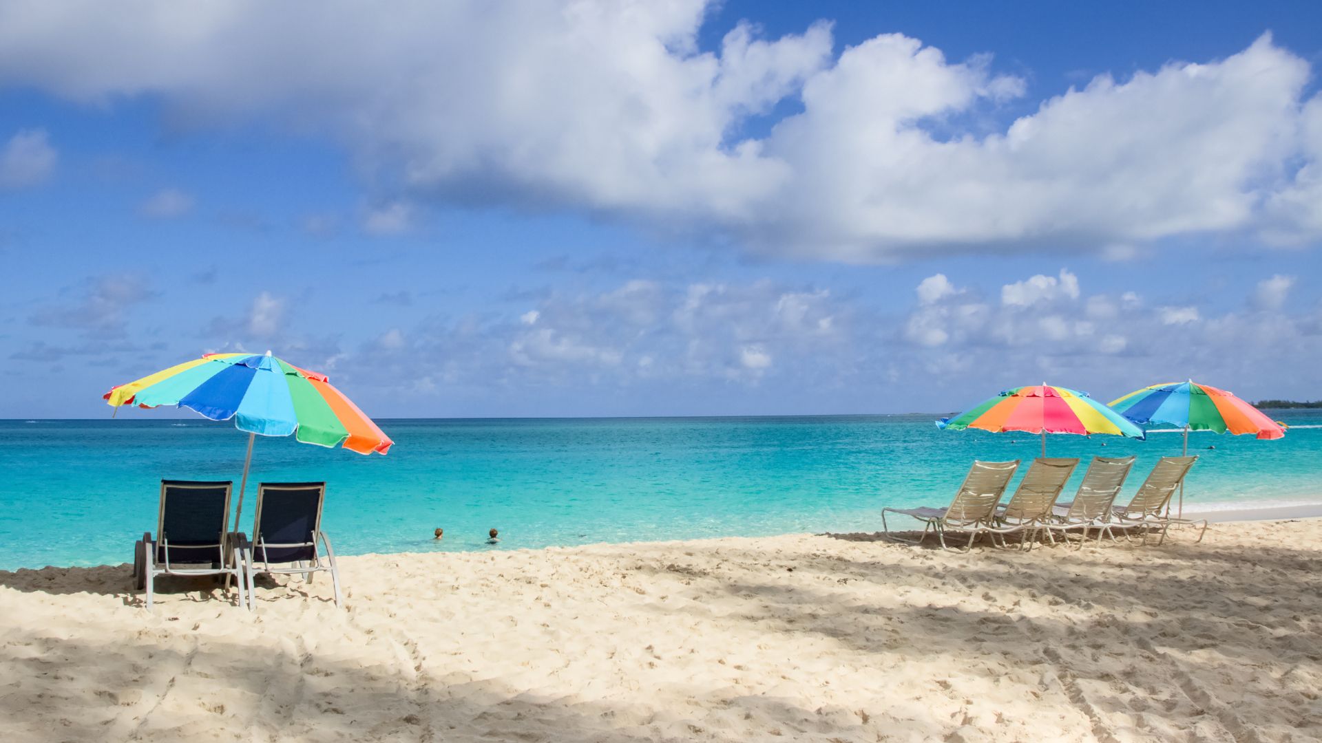 A white sand beach with turquoise water features two multi-colored beach umbrellas, several lounge chairs, and people swimming in the ocean.