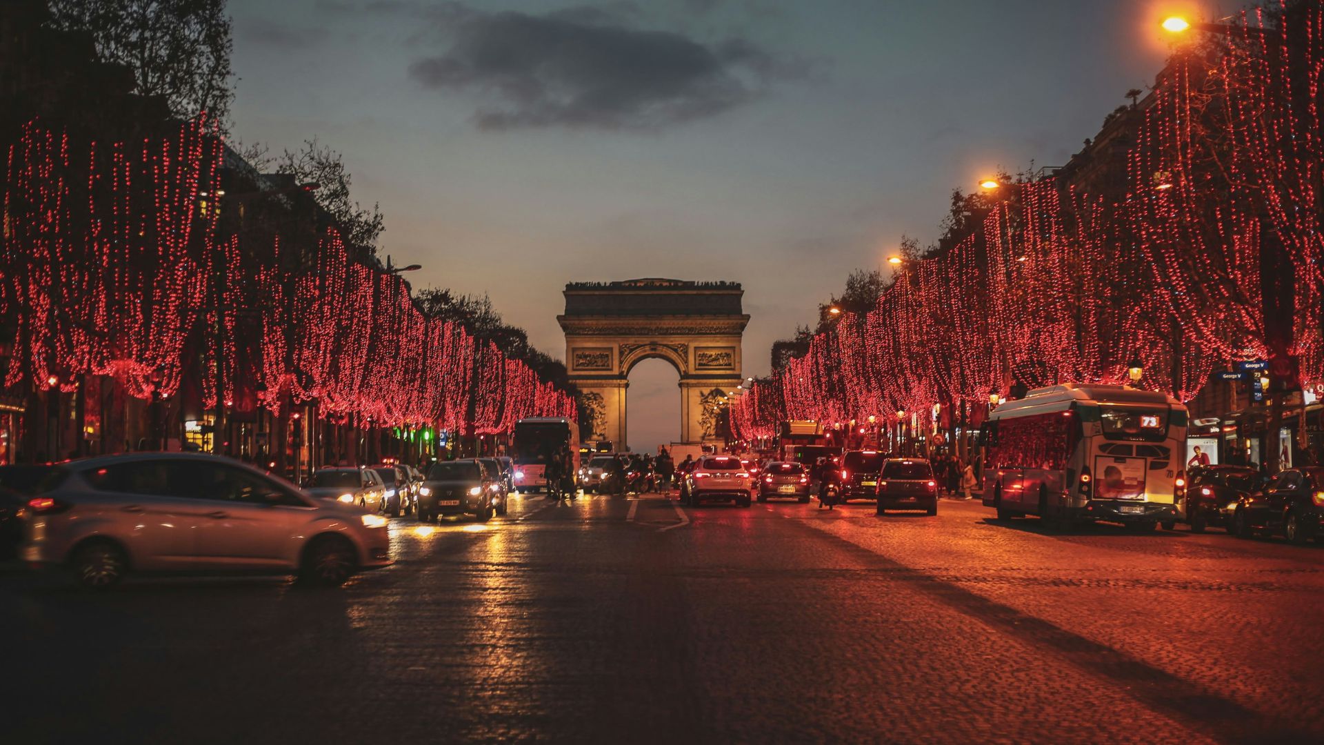 The Avenue des Champs-Élysées in Paris is seen at dusk with heavy traffic and trees illuminated by red Christmas lights, leading the eye to the Arc de Triomphe in the background.