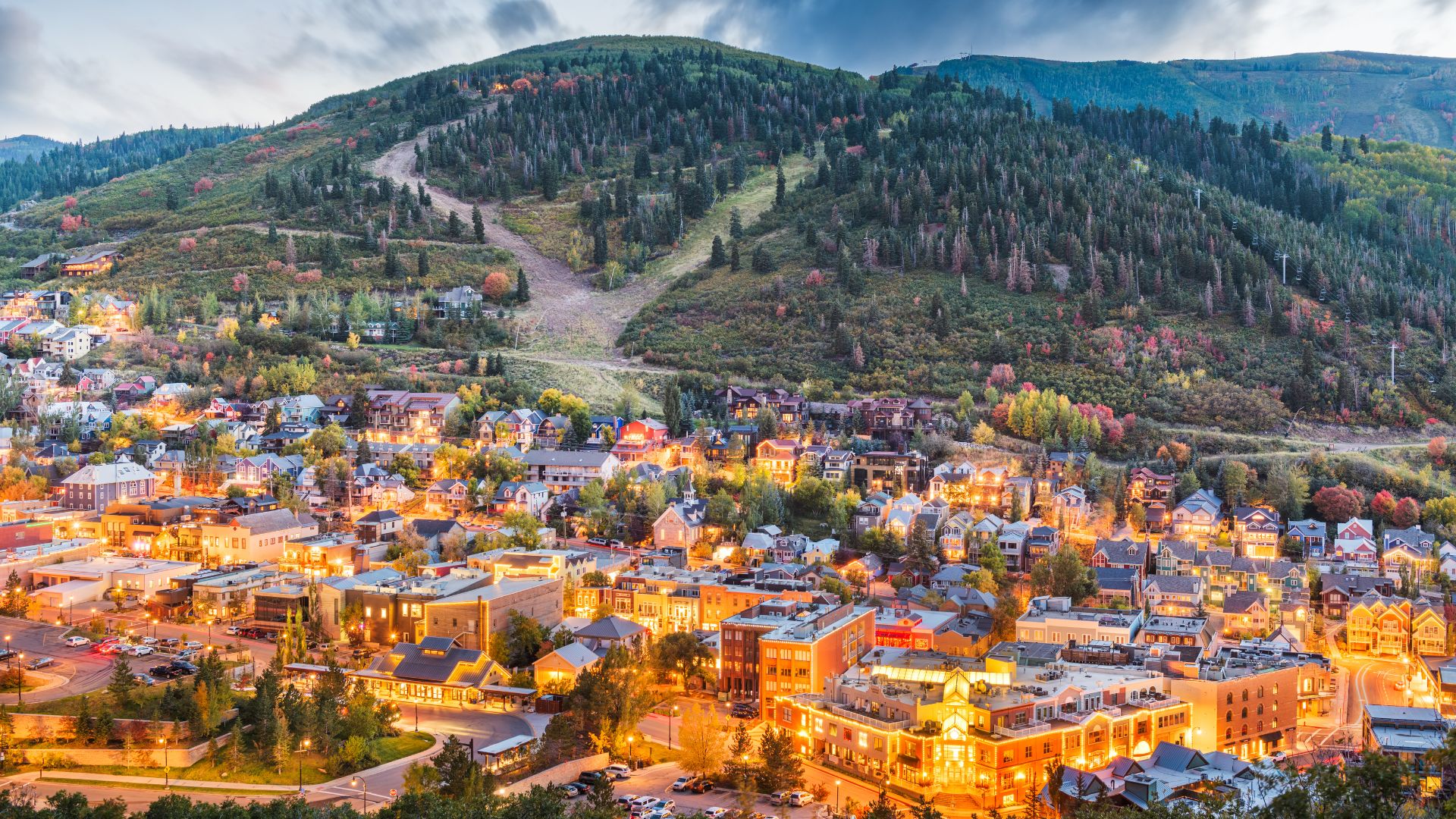 An aerial view of Park City, Utah at dusk, showing a lit-up town at the base of a forested mountain with a ski slope.