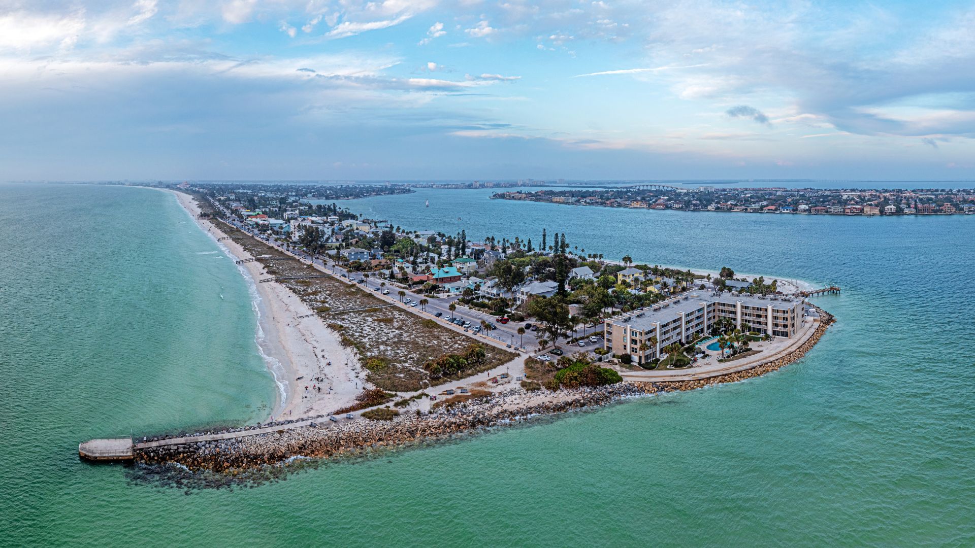 An aerial daytime photograph of the narrow, sandy peninsula of Pass-a-Grille Beach in St. Pete Beach, Florida, showing low-rise buildings nestled between the turquoise Gulf of Mexico and the bay, with a rock jetty in the foreground.