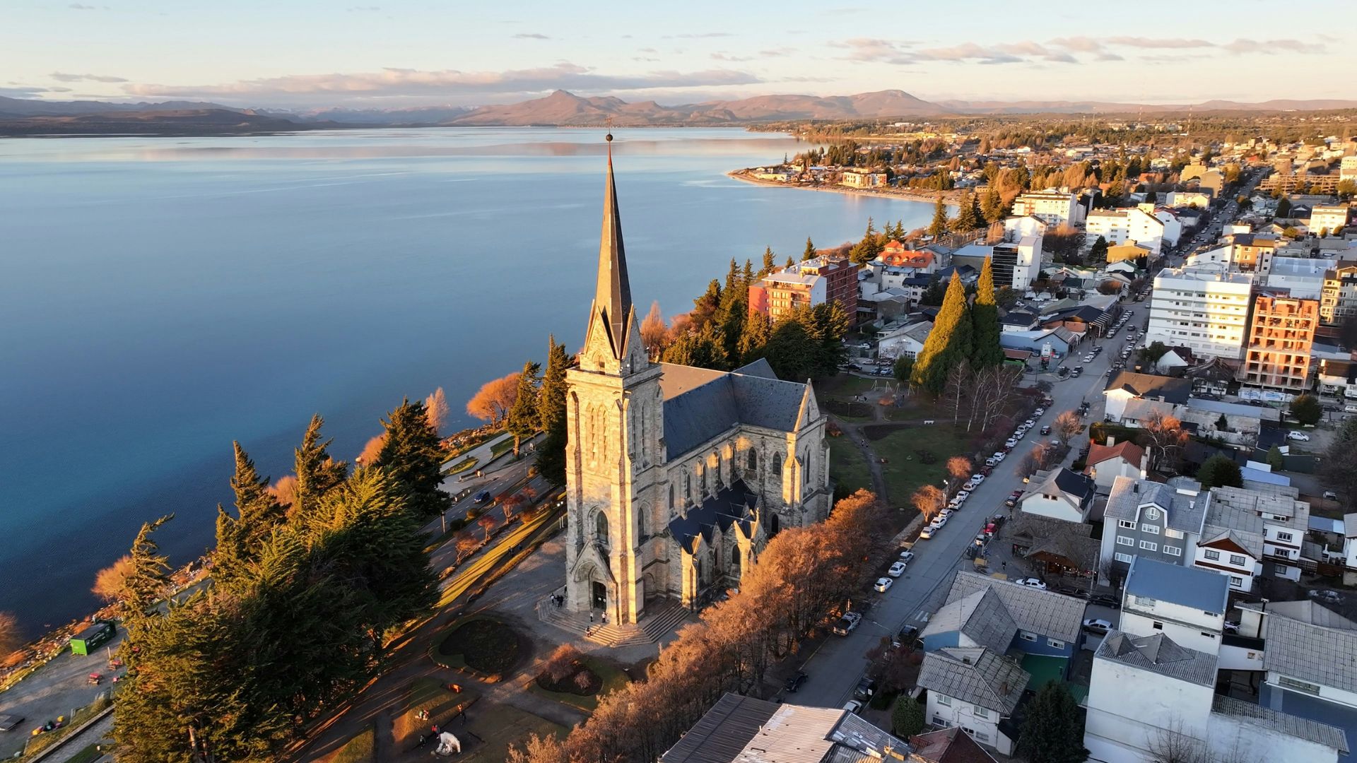 An elevated, sunlit view of the neo-Gothic Cathedral of Our Lady of Nahuel Huapi and the surrounding town of Bariloche on the shores of Lake Nahuel Huapi, with mountains in the background.