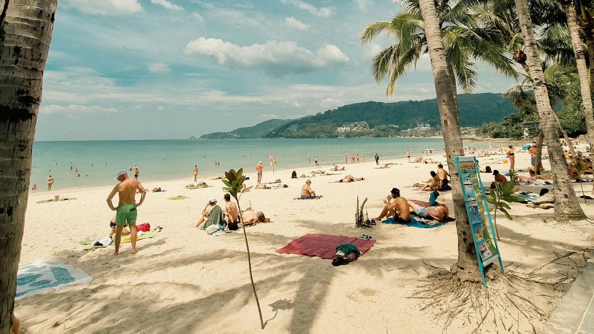 People on a sandy beach with palm trees and hills in the background on a sunny day
