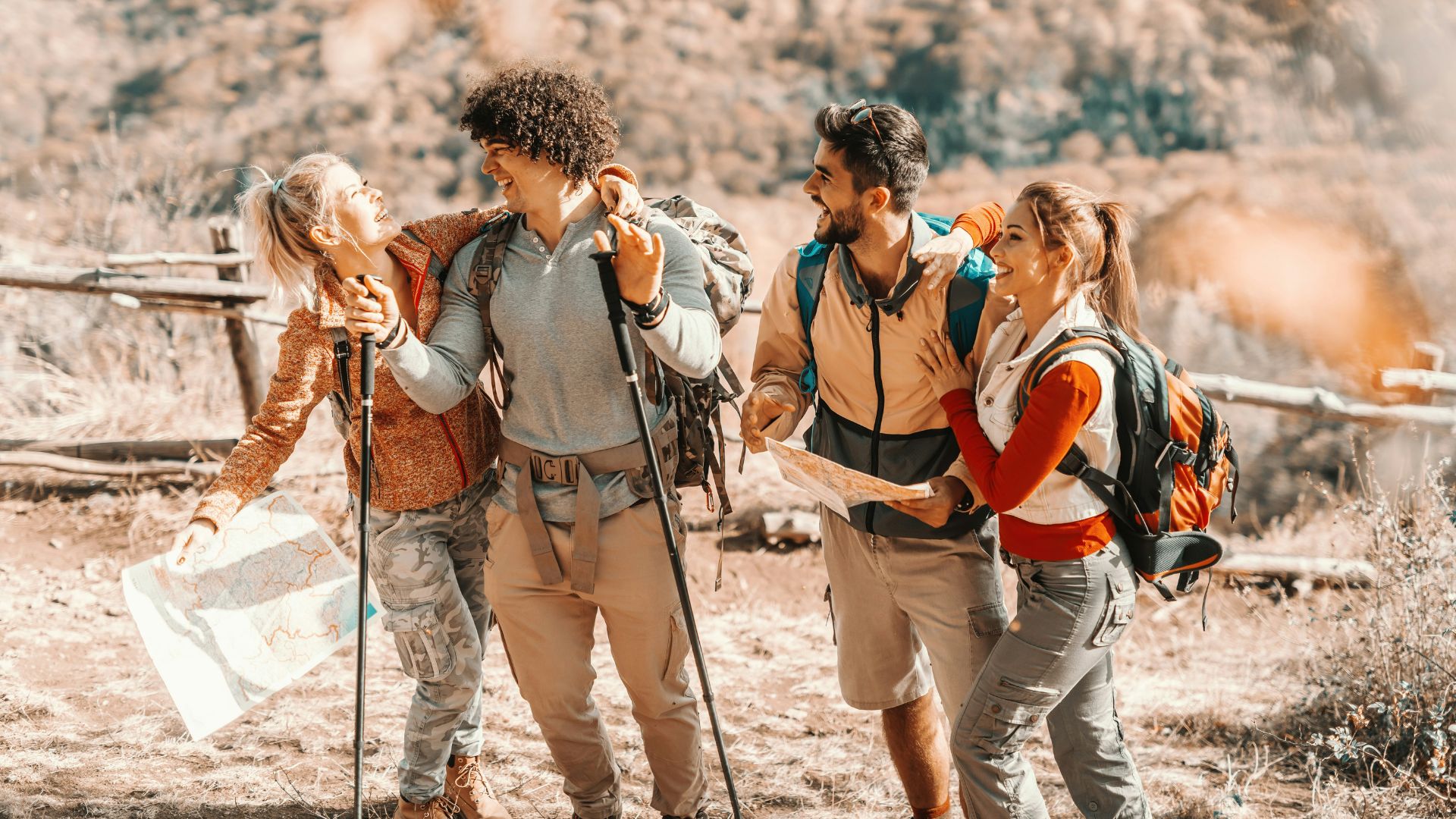 A group of four happy hikers with backpacks and trekking poles consult a map on a dry, grassy mountain trail.