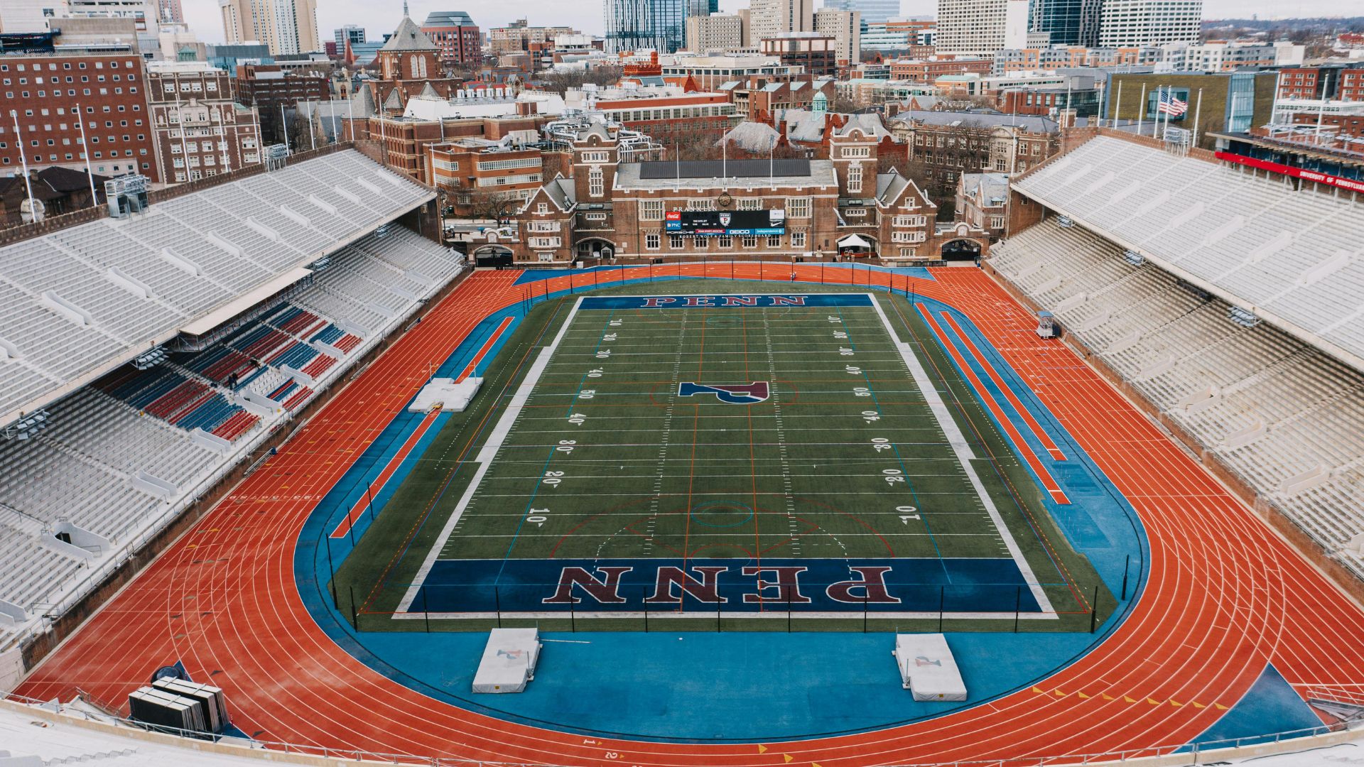 An aerial view of the Franklin Field stadium and track, with the 'PENN' logo painted on the blue end zones and the Philadelphia skyline visible in the background.