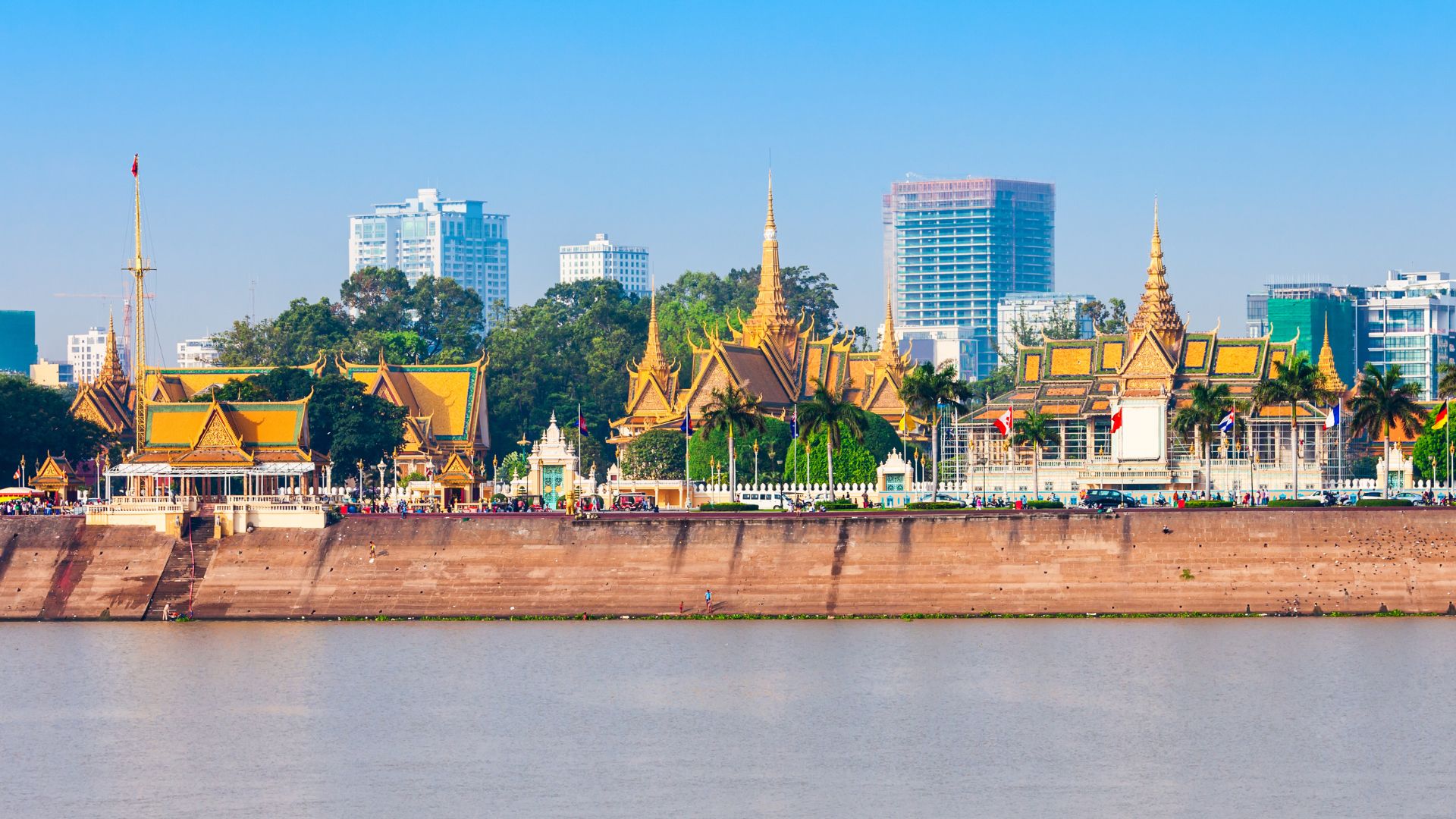 A view of the ornate Royal Palace complex in Phnom Penh, Cambodia, situated by a river with modern city buildings in the background.