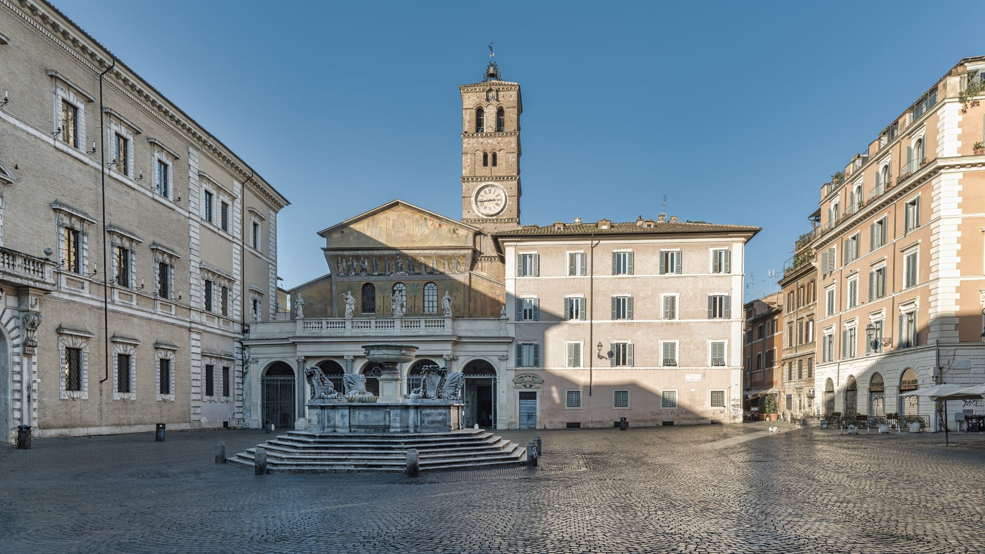 A wide view of a cobblestone square in Rome with an octagonal fountain in the foreground, facing the historic stone facade of the Basilica of Santa Maria in Trastevere and its tall clock tower.